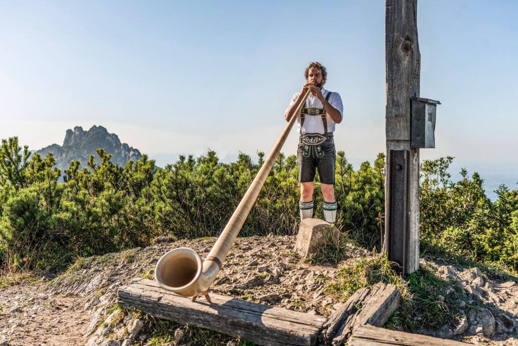 Hornist Christian Loferer bläst an einem Gipfelkreuz in sein Alphorn.