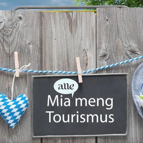 Ein Schild mit der Aufschrit "Mia alle meng Tourismus" hängt an einer Kordel, daneben hängen eine Brezel und ein Hut.