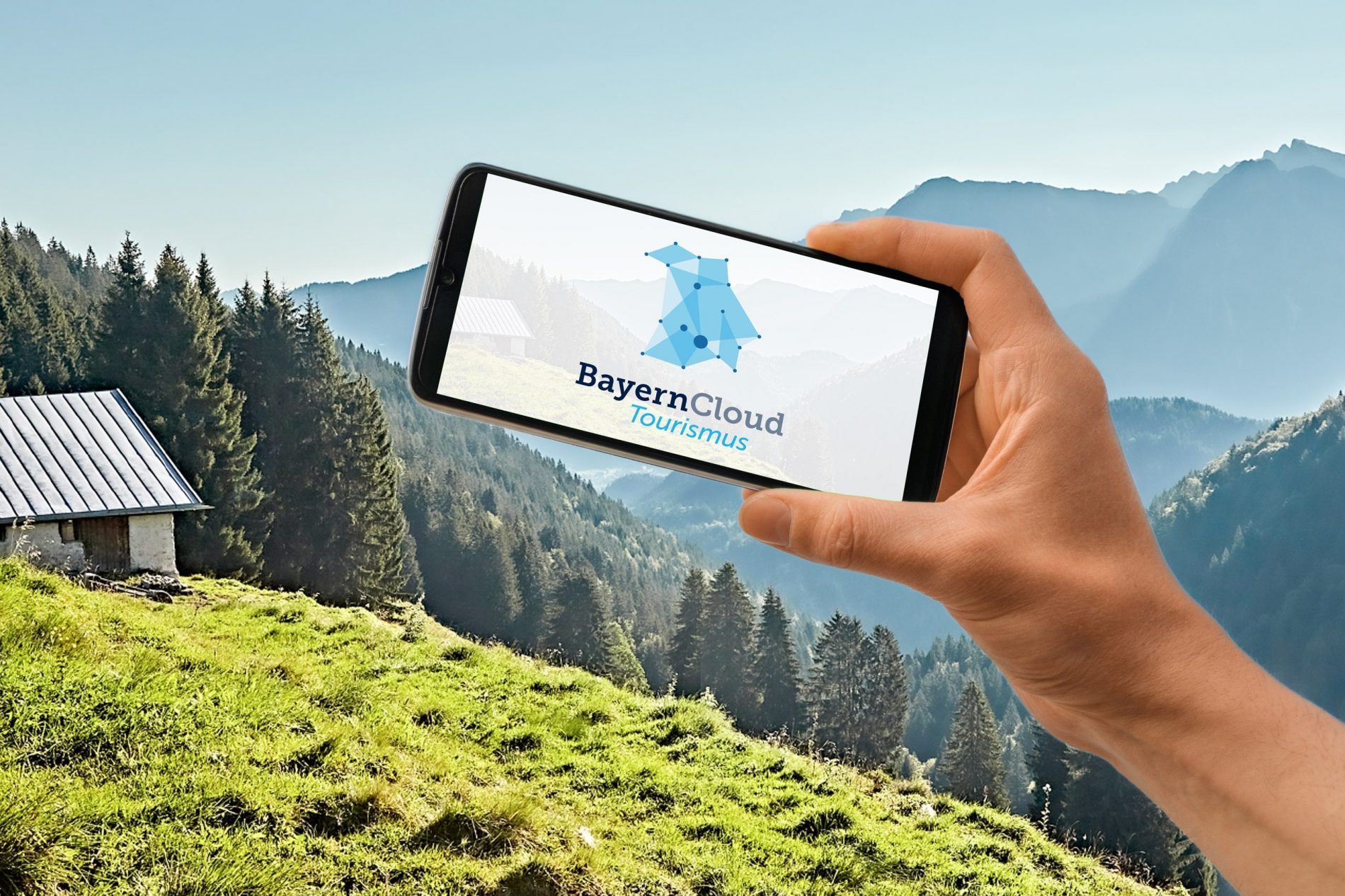BayernCloud Tourismus Smartphone mit dem Logo der BayernCloud Tourismus auf dem Bildschirm, im Hintergrund sind Berge und Wiesen zu sehen.