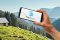 Smartphone mit dem Logo der BayernCloud Tourismus auf dem Bildschirm, im Hintergrund sind Berge und Wiesen zu sehen.