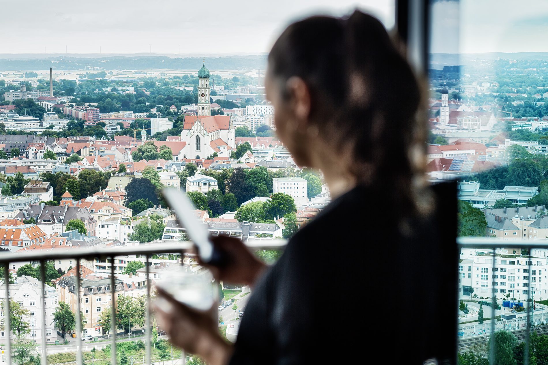 Augsburg, Dorint Hotel am Kongress, MICE, Shooting Sommer 2021 Frau mit Smartphone und Kaffeetasse in der Hand vor einem Fenster mit Ausblick auf die Stadt Augsburg