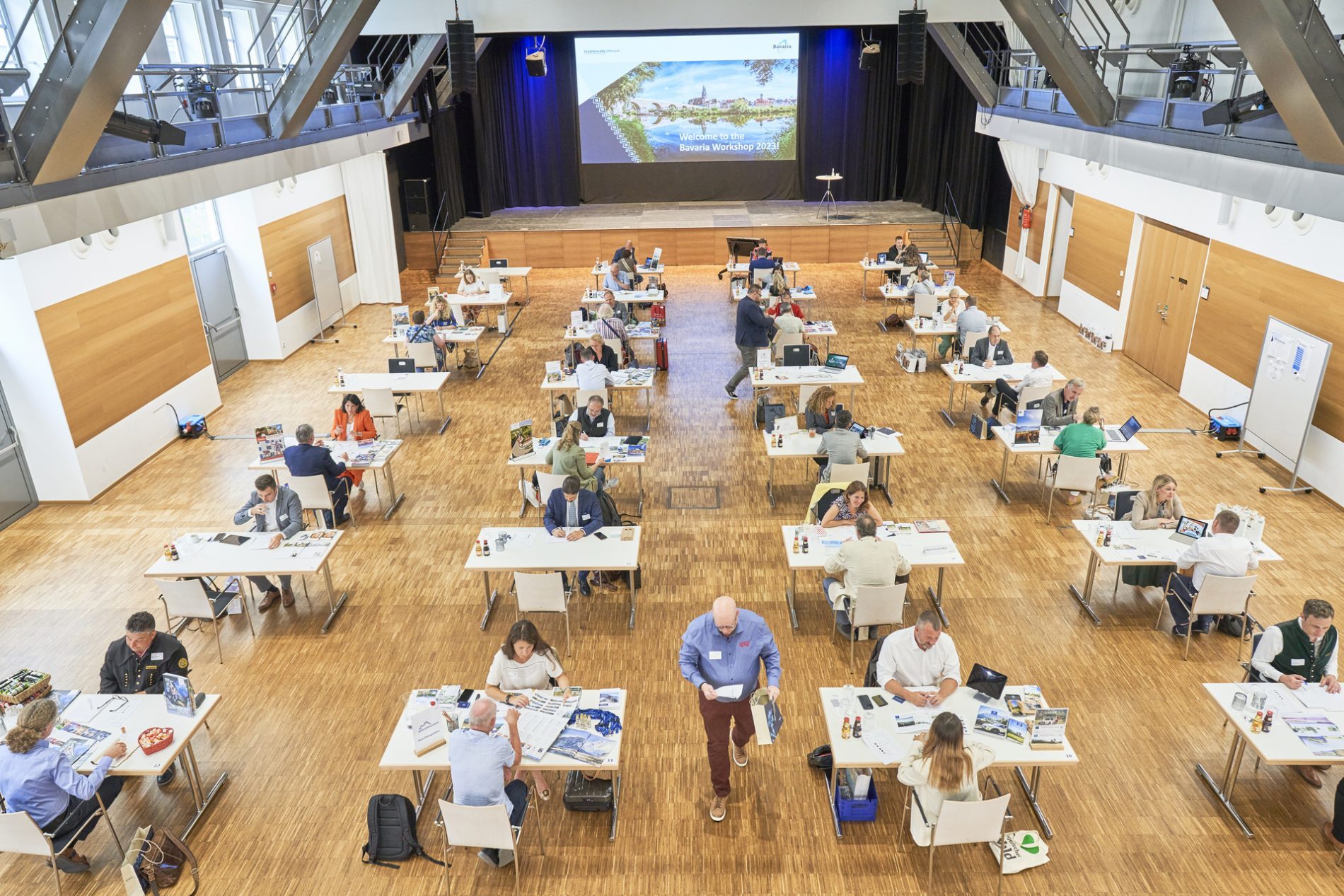 Heller Veranstaltungsraum mit vielen Tischen und Teilnehmern; Bühne mit Bildschirm „Welcome to the Bavaria Workshop 2023“.