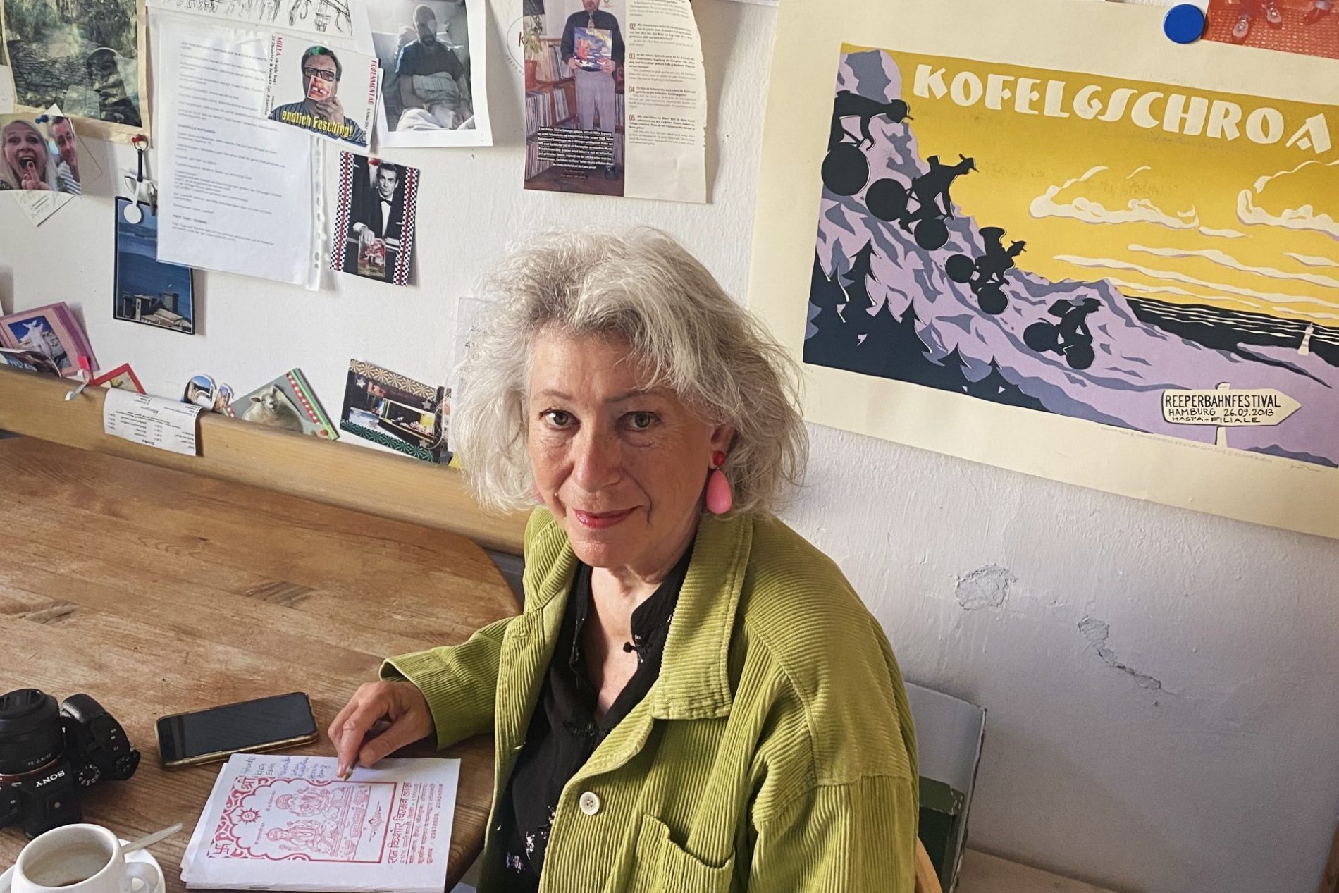 Angelika Jakob Porträt einer älteren Frau mit grauem Haar in grüner Jacke, an Tisch sitzend; Wand voller Poster und Collagen im Hintergrund.