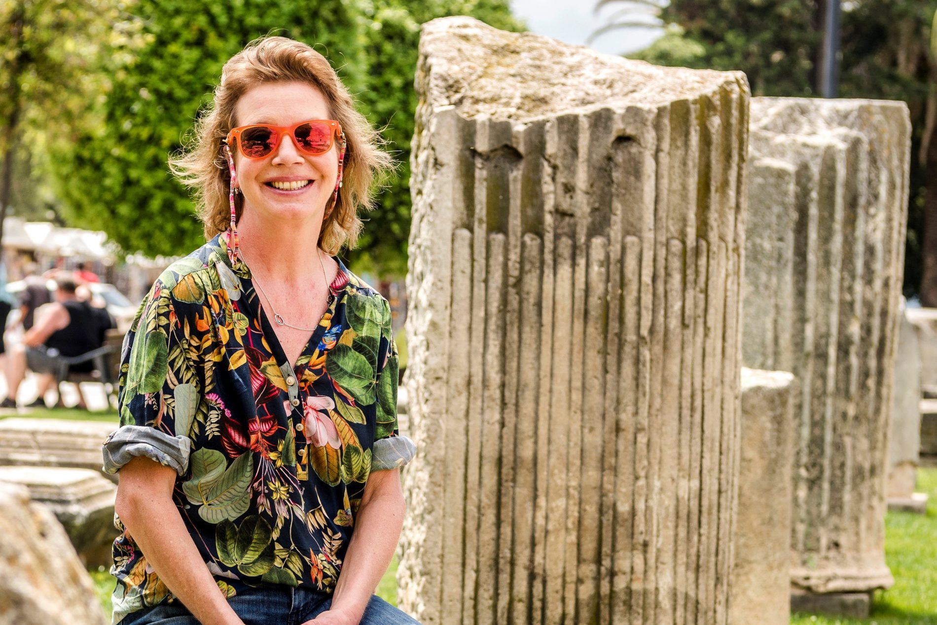 Anja Keul_2 Anja Keul sitzt im Freien neben antiken Stein-Säulen, trägt buntes tropisches Hemd und orange Sonnenbrille, lächelt.