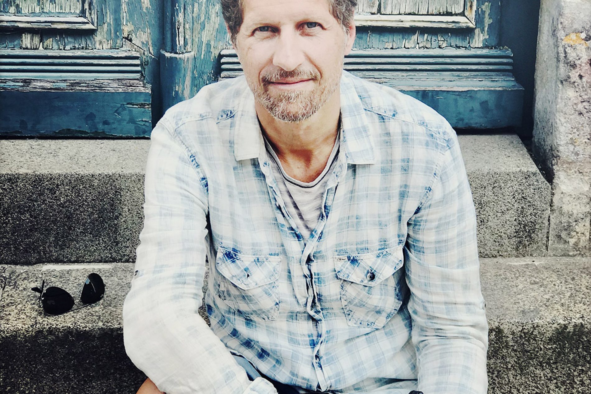 Bernhard_Huber Mann sitzt auf Stufen vor abgeblätterter blauer Tür, trägt helles Karohemd und hält eine Vintage-Kamera.