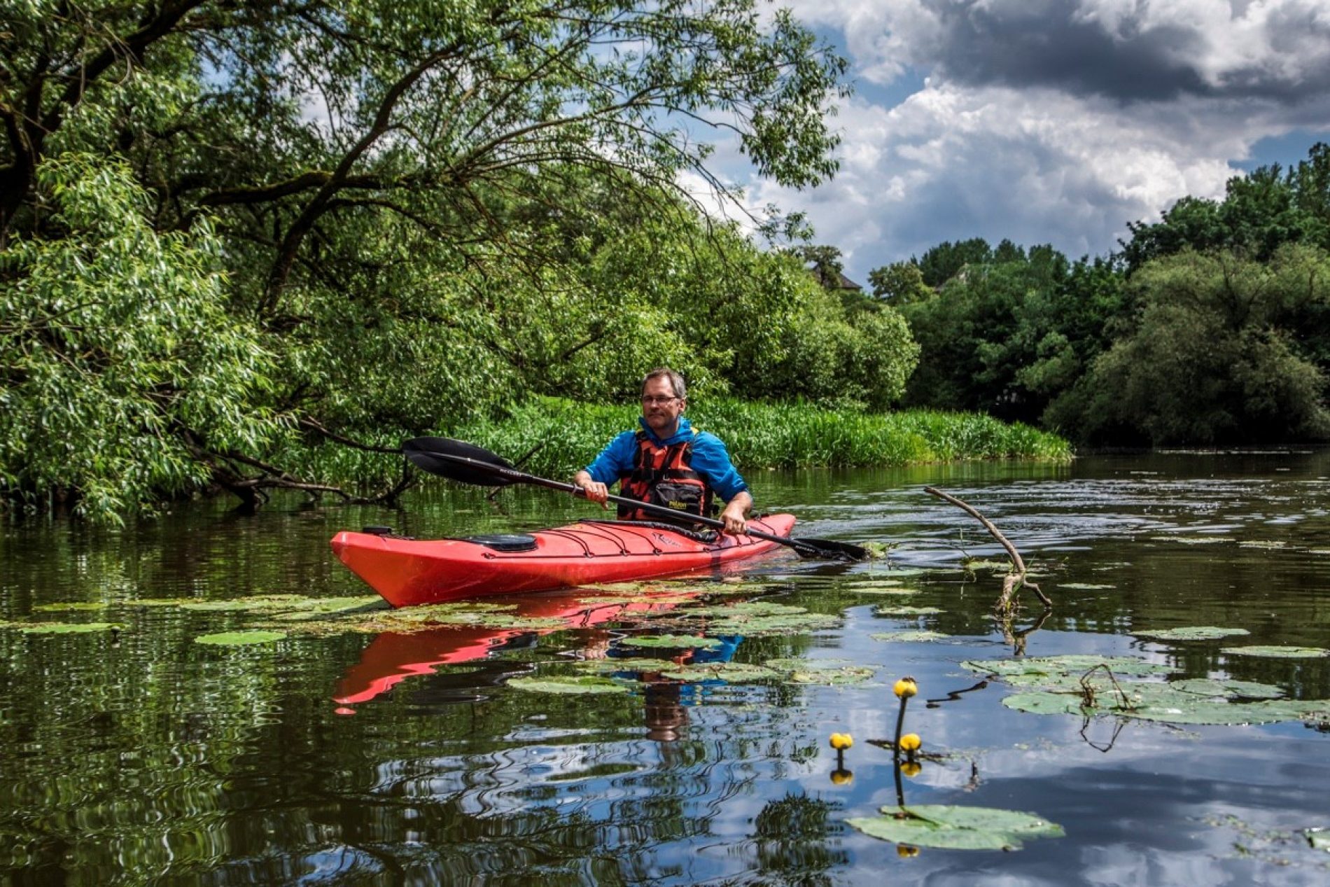 Christian_Haas Christian Haas paddelt in rotem Kajak auf ruhigem Fluss mit Seerosen, grünem Ufer und Wolkenhimmel.
