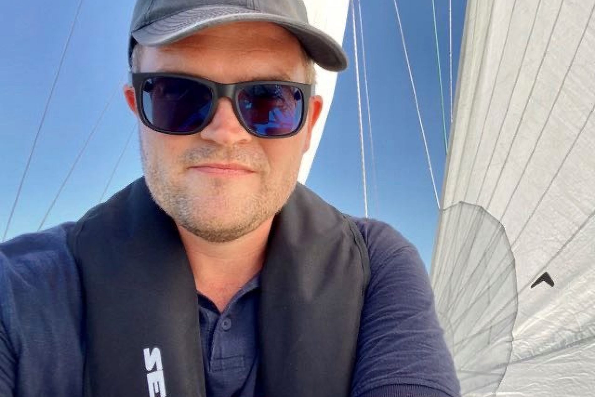 Florian-Trykowski Florian Trykowski mit Baseballkappe, Sonnenbrille und Rettungsweste auf Segelboot bei sonnigem Himmel.