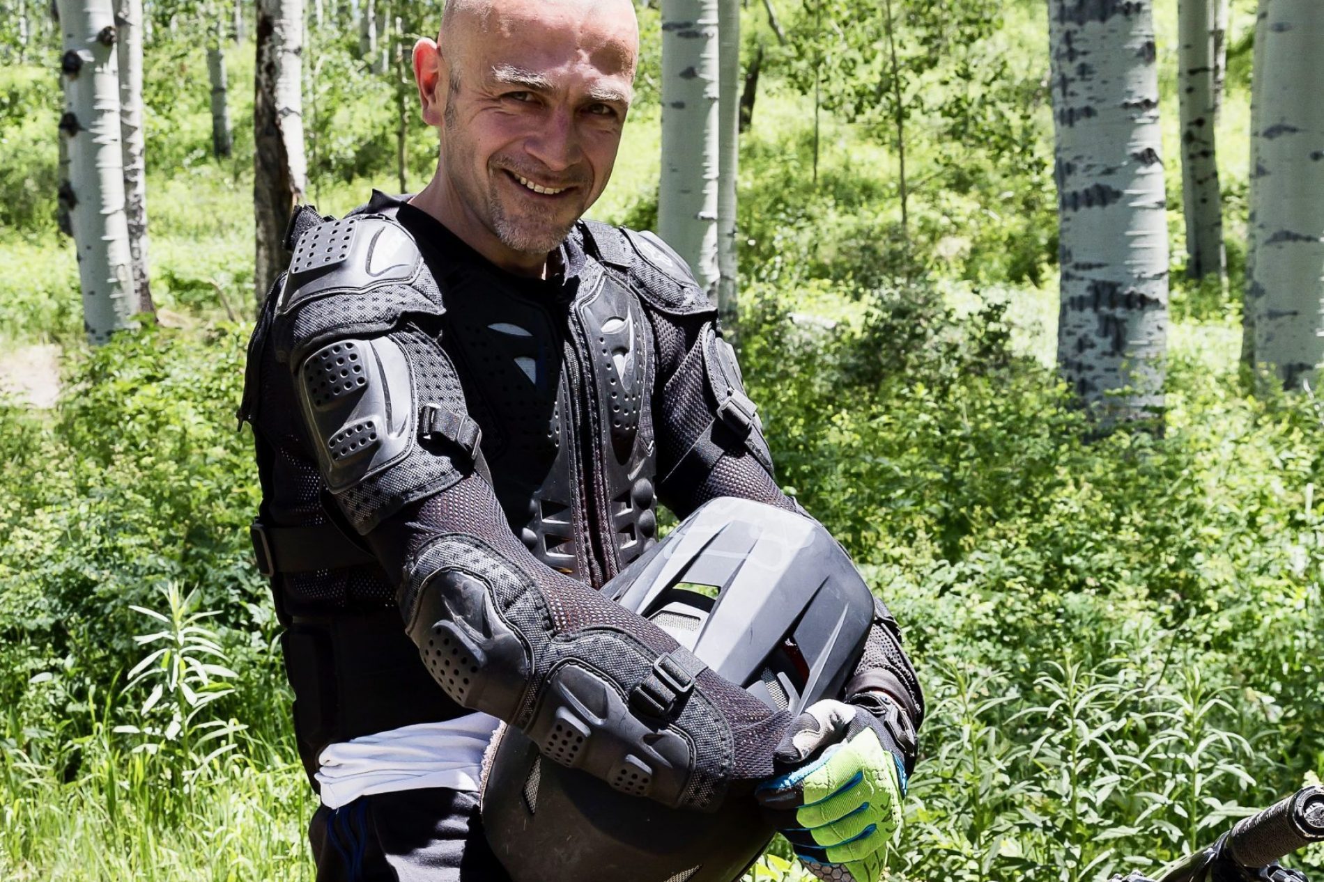 Flori in Aspen 2017 Lächelnder Mann in Schutzkleidung mit Brustplatten hält Helm, neben gelbem Mountainbike im Birkenwald.