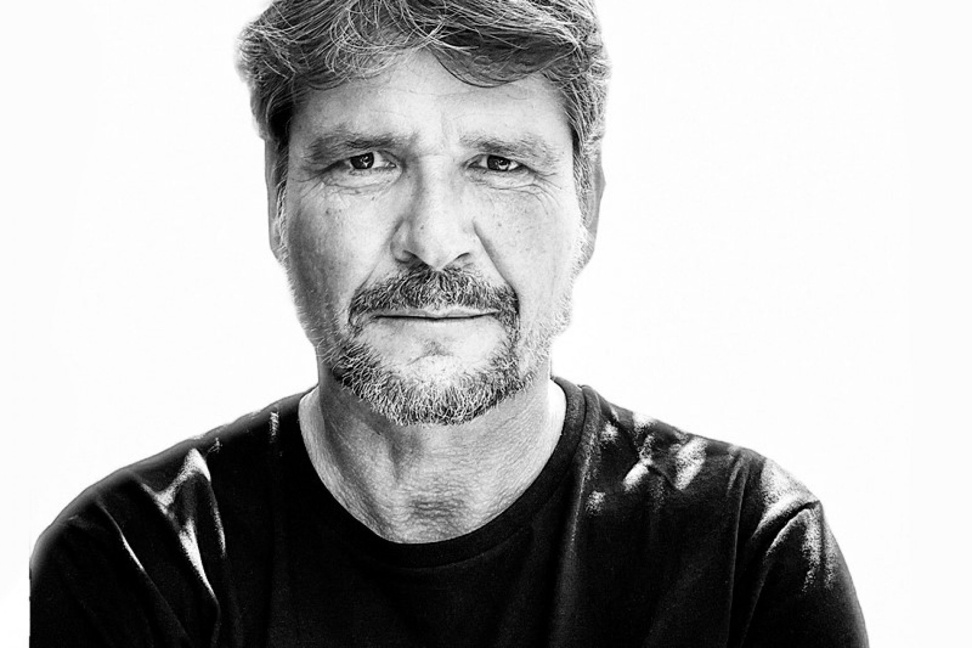 Frank_Heuer Porträt von Frank Heuer in Schwarz-Weiß: mittlerer Alter Mann mit grauem Bart, dunkles T-Shirt, Arme leicht verschränkt.