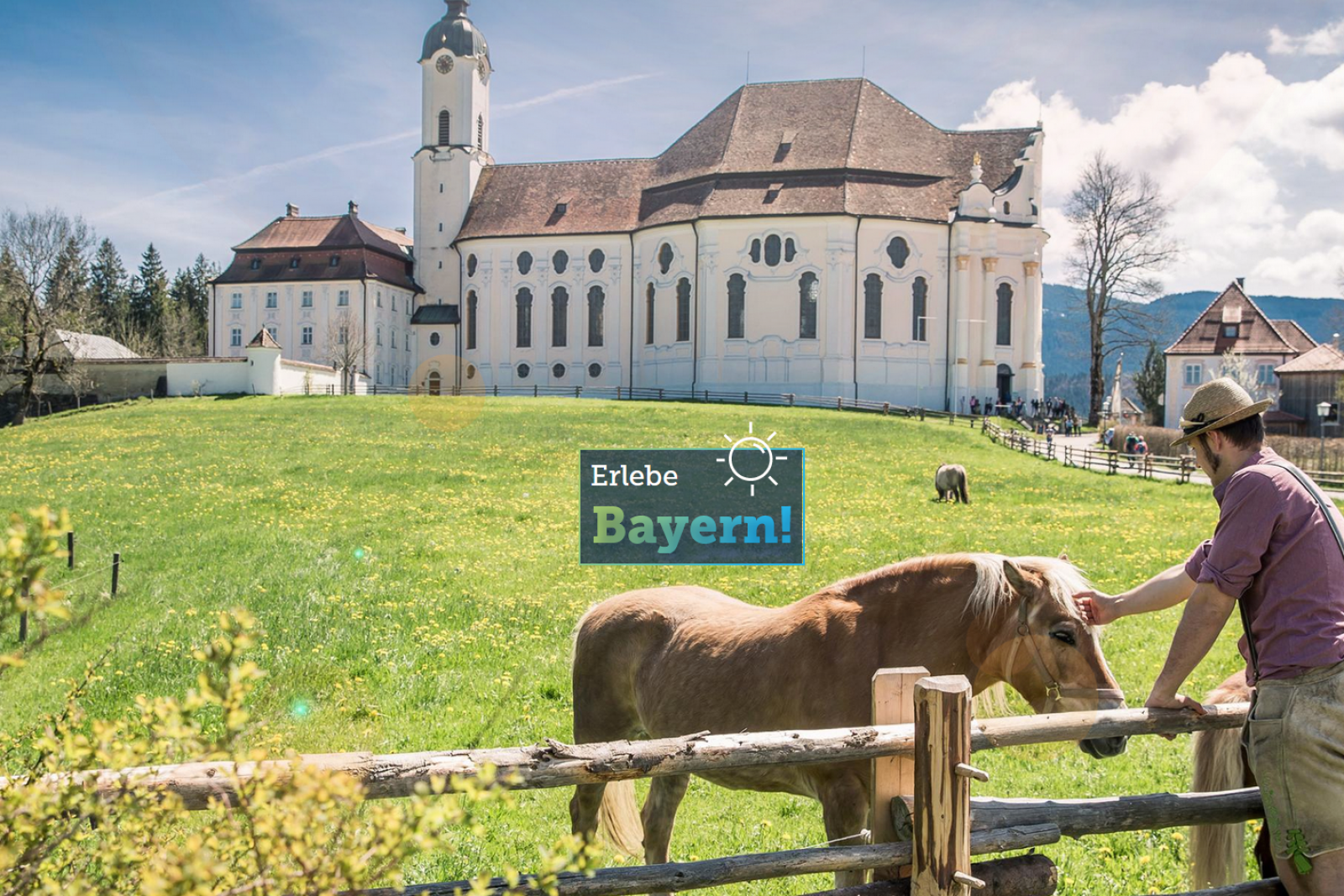 Weiße Klosteranlage auf grünem Feld, Mann streichelt ein Pferd hinter Holzzaun; Wald, Berge und blauer Himmel im Hintergrund