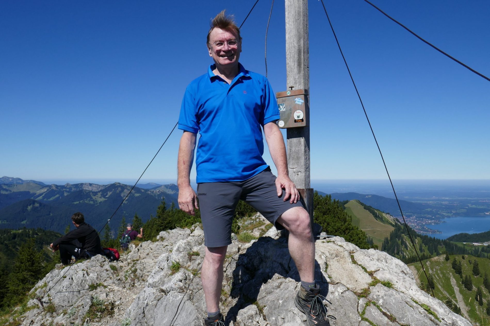 Markus Stein Markus Stein im blauen Polo steht auf felsigem Gipfel neben einem Holzpfosten mit Kabeln, Blick auf Berge und See.