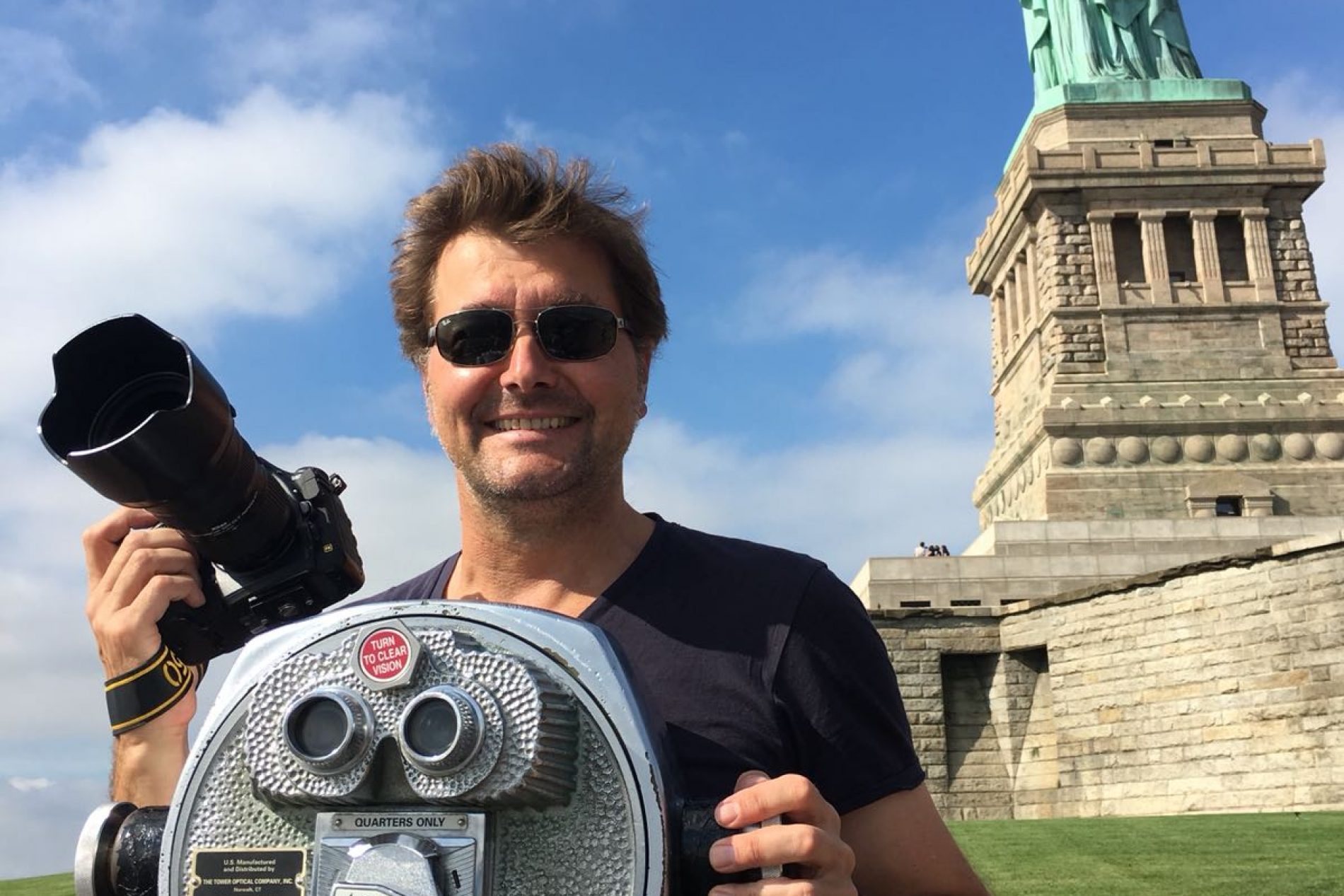 Norbert Eisele-Hein Mann mit Sonnenbrille und schwarzem T-Shirt posiert neben Münzfernrohr; Statue of Liberty-Replik im Hintergrund.