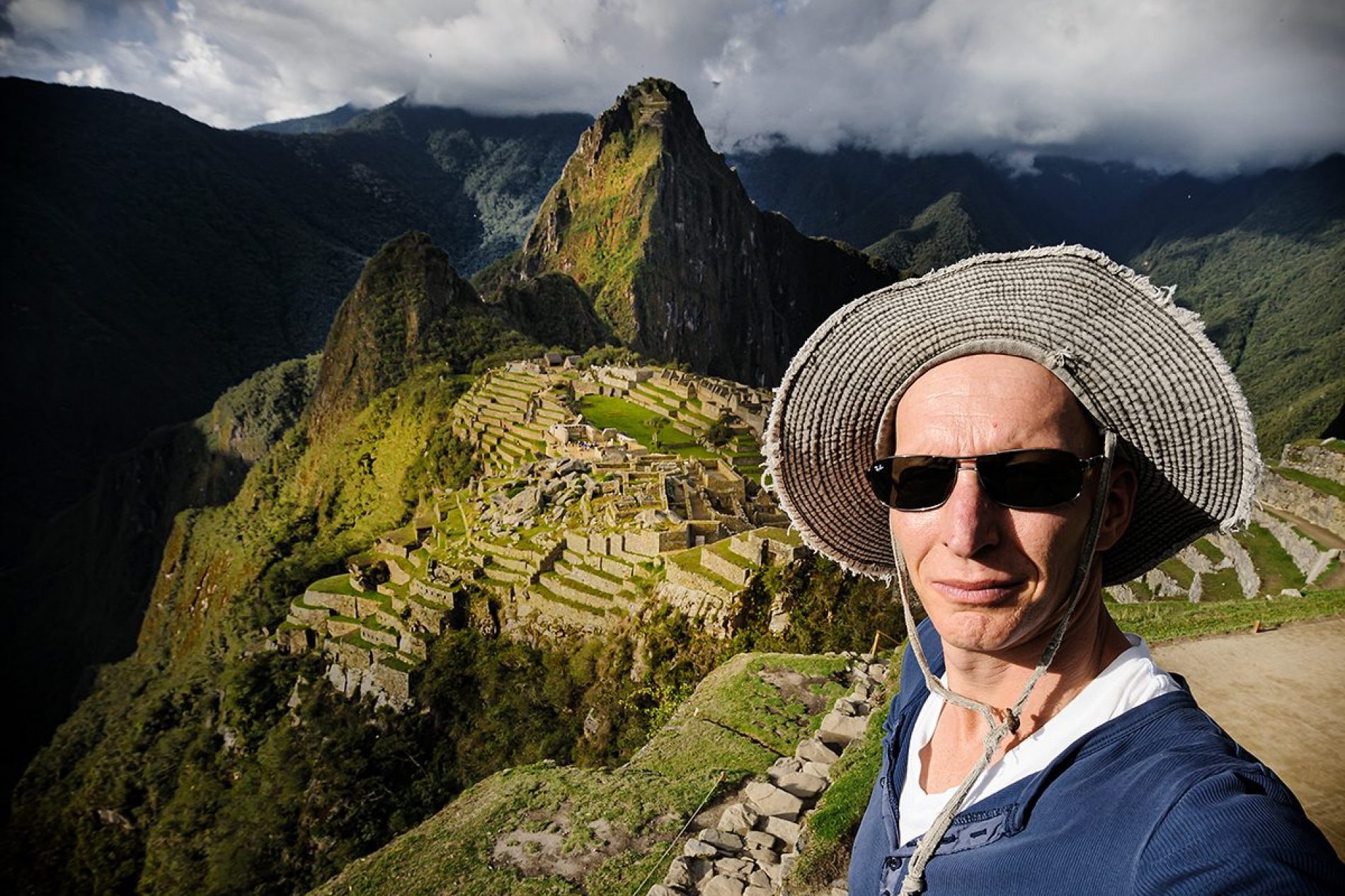 Thomas_Linkel Mann mit Strohhut und Sonnenbrille posiert vor den Terrassen von Machu Picchu in bergiger Landschaft.