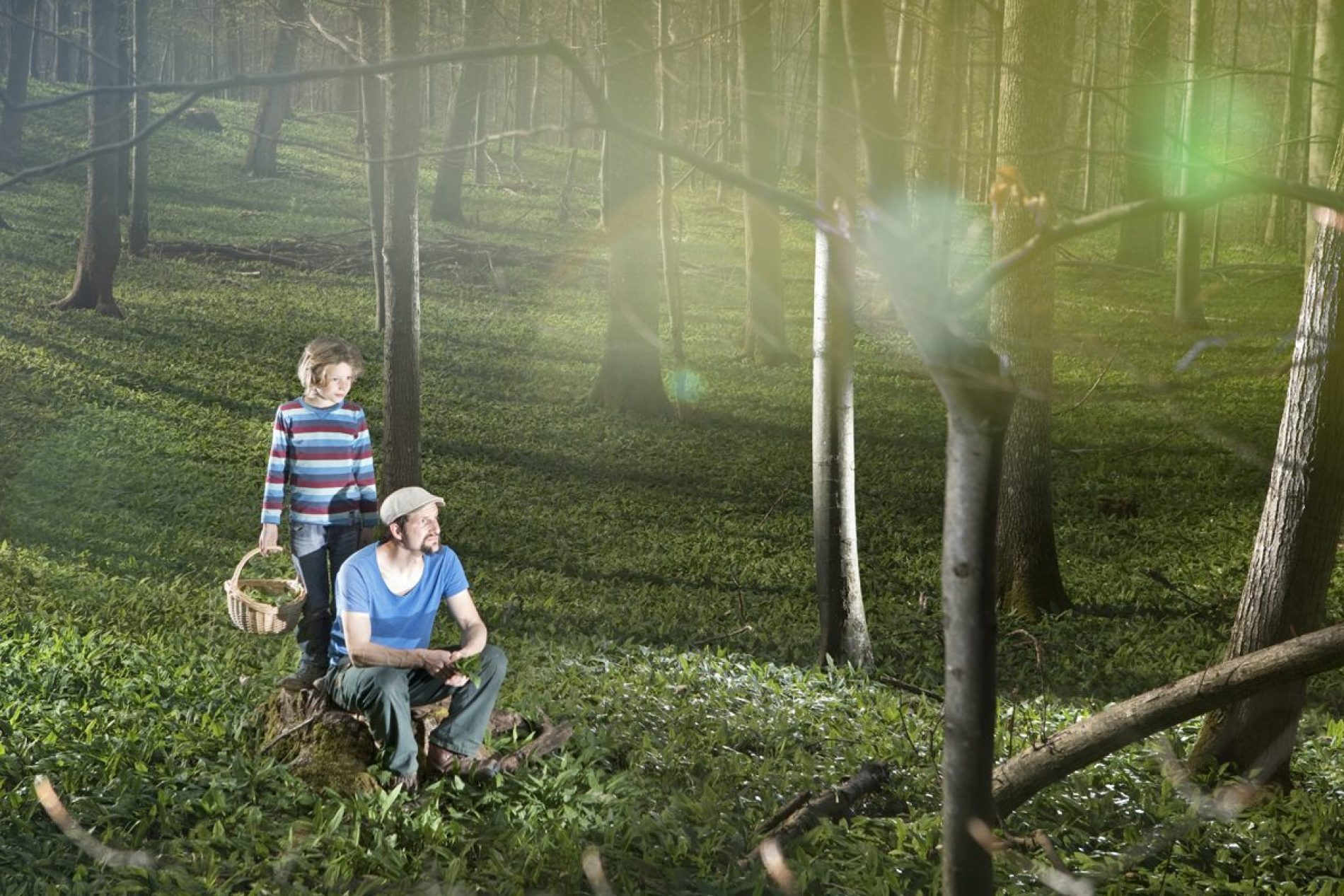 Tobias_Gerber Zwei Personen im Wald: Ein Mann sitzt auf einer Baumwurzel, daneben steht ein Junge mit Korb; Lichtstrahlen durch Bäume.