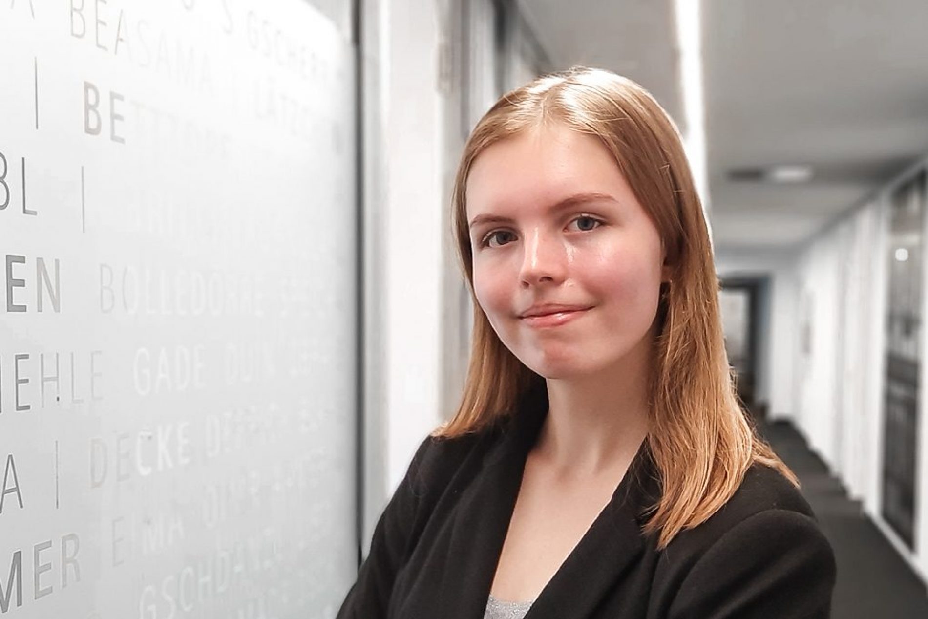 Yevheniia Lehkodymova Porträt einer jungen Frau mit hellbraunen Haaren in schwarzer Jacke, im Flur neben einer beschrifteten Glaswand.