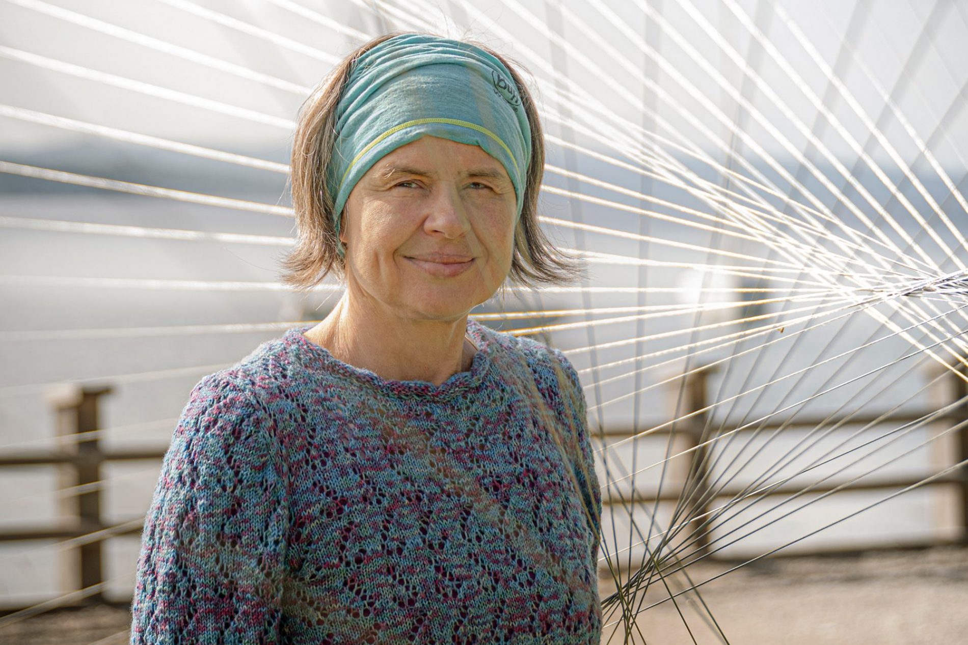 karin-lochner Portrait einer Frau mit türkisgrüner Kopfbedeckung und buntem Strickpullover, umgeben von feinen Fasersträngen im Freien