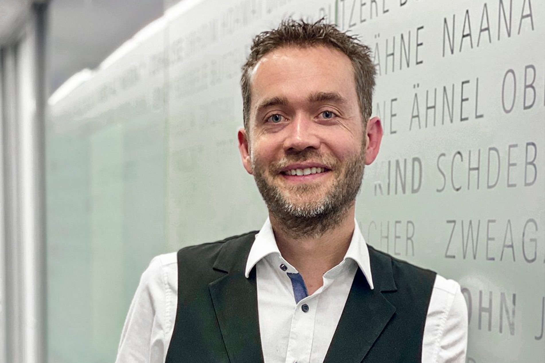 Andreas Höckinger Porträt eines lächelnden Mannes in weißem Hemd und schwarzer Weste vor Glaswand mit Schriftzügen; Bezug: Andreas Höckinger