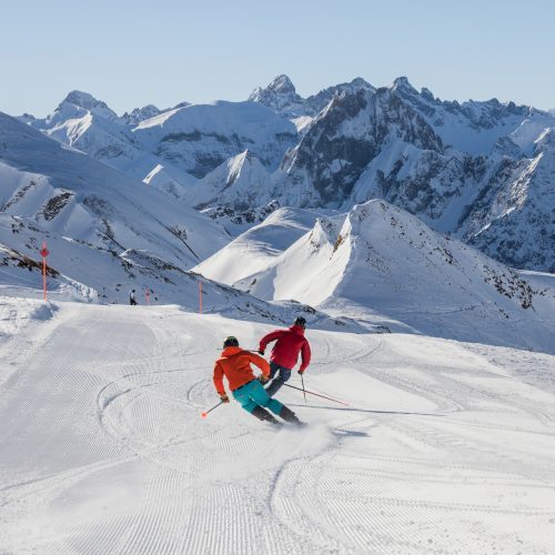 Zwei Personen fahren bei schönstem Wetter Ski, im Hintergrund ist eine Bergkette zu sehen.