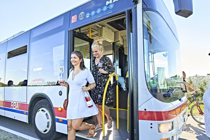 Zwei Frauen steigen aus einem Bus des Öffentlichen Personennahverkehrs. Sie haben Weingläser in der Hand und das Wetter ist sommerlich.