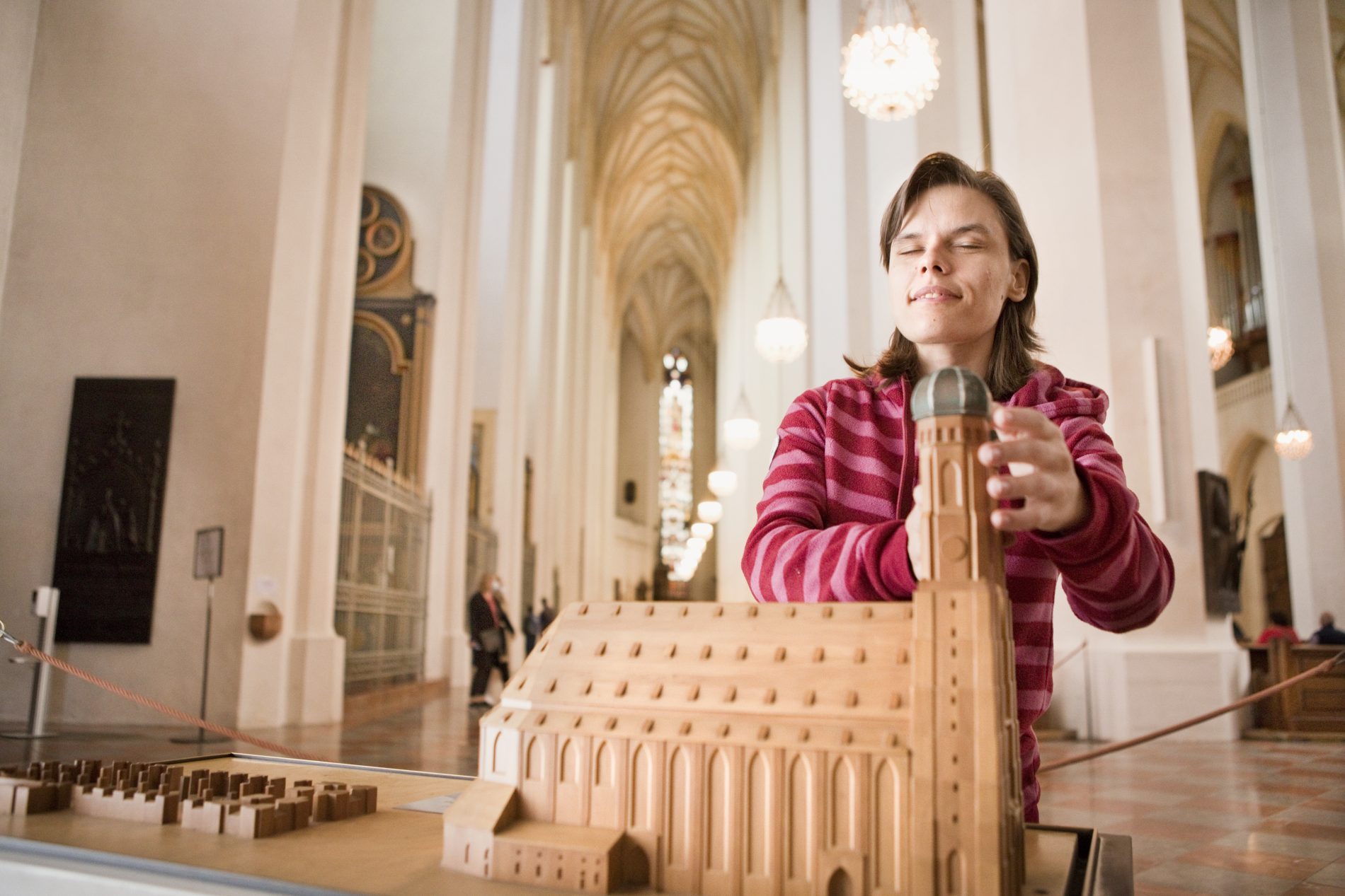 Frau in rotem Hoodie baut in einer Hallenkathedrale ein Holzkatedral-Modell; Hintergrund mit Besuchern.