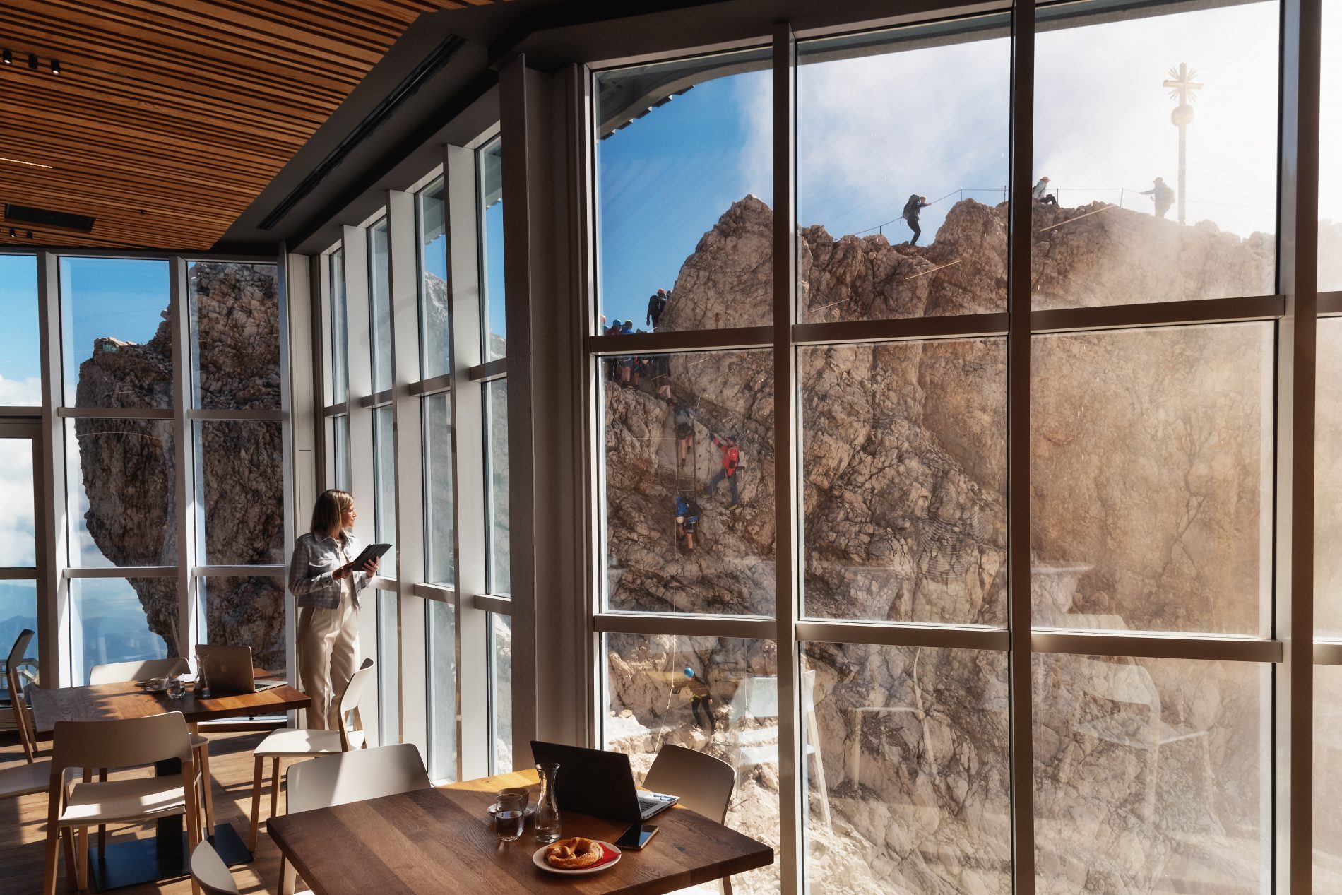 Innenraum mit gro&szlig;en Glasscheiben, Tischen und Laptop; drau&szlig;en klettern Bergsteiger an felsigem Gipfel.