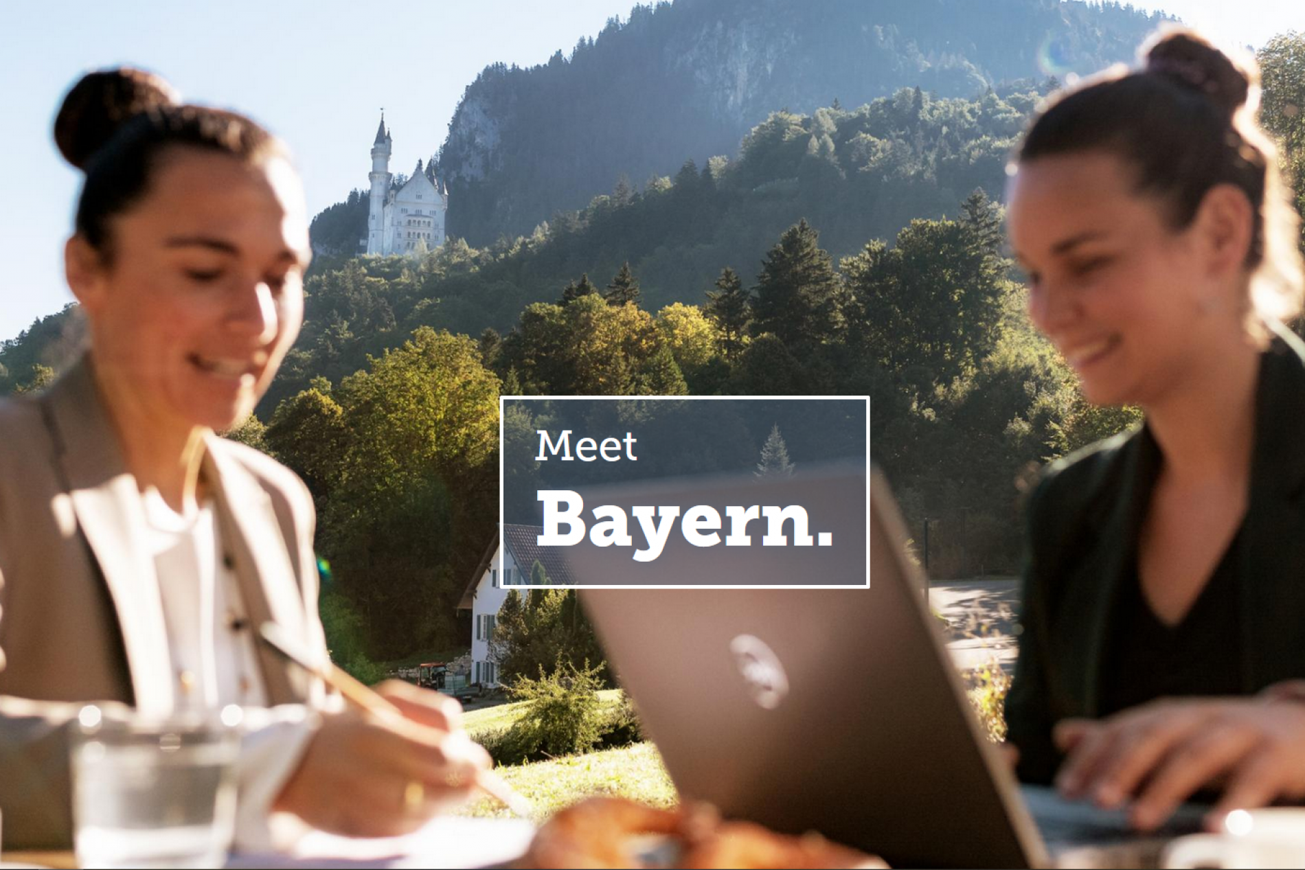 meet.bayern-1 Zwei Frauen arbeiten draußen mit Laptop vor Berglandschaft und Schloss im Hintergrund, Sponsored Tipp Meet Bayern.