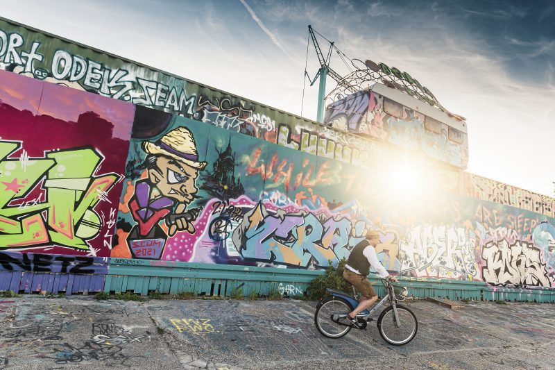 Mauer mit Graffiti. Davor fährt ein Mann auf einem Fahrrad vorbei.