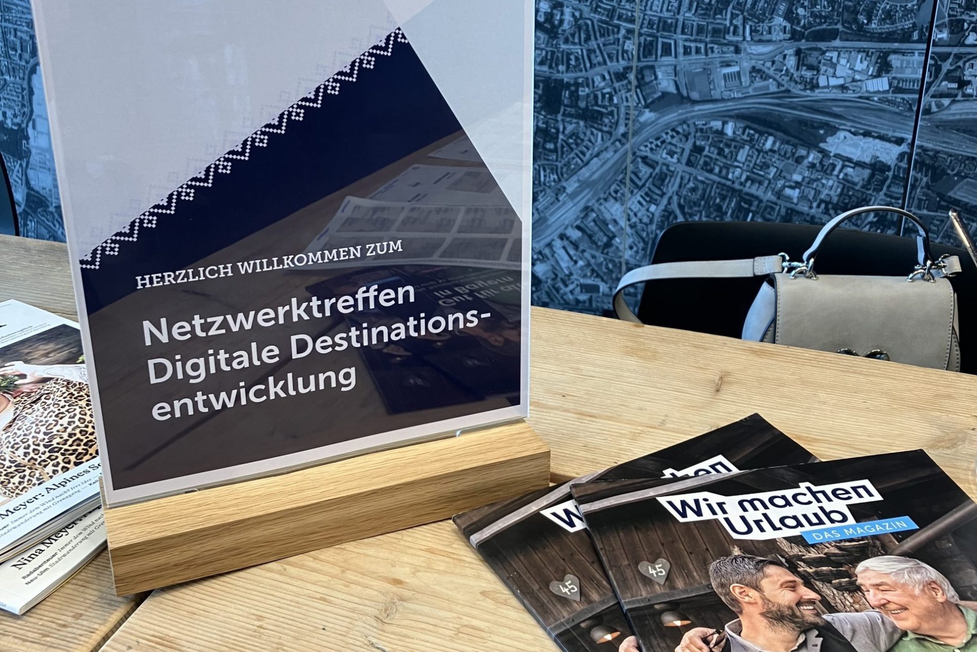 Netzwerktreffen Digitale Destinationsentwicklung Banner mit Bayern-Logo und Text Netzwerktreffen Digitale Destinationsentwicklung vor einem Kartenausschnitt; Holztisch, Zeitschriften daneben.