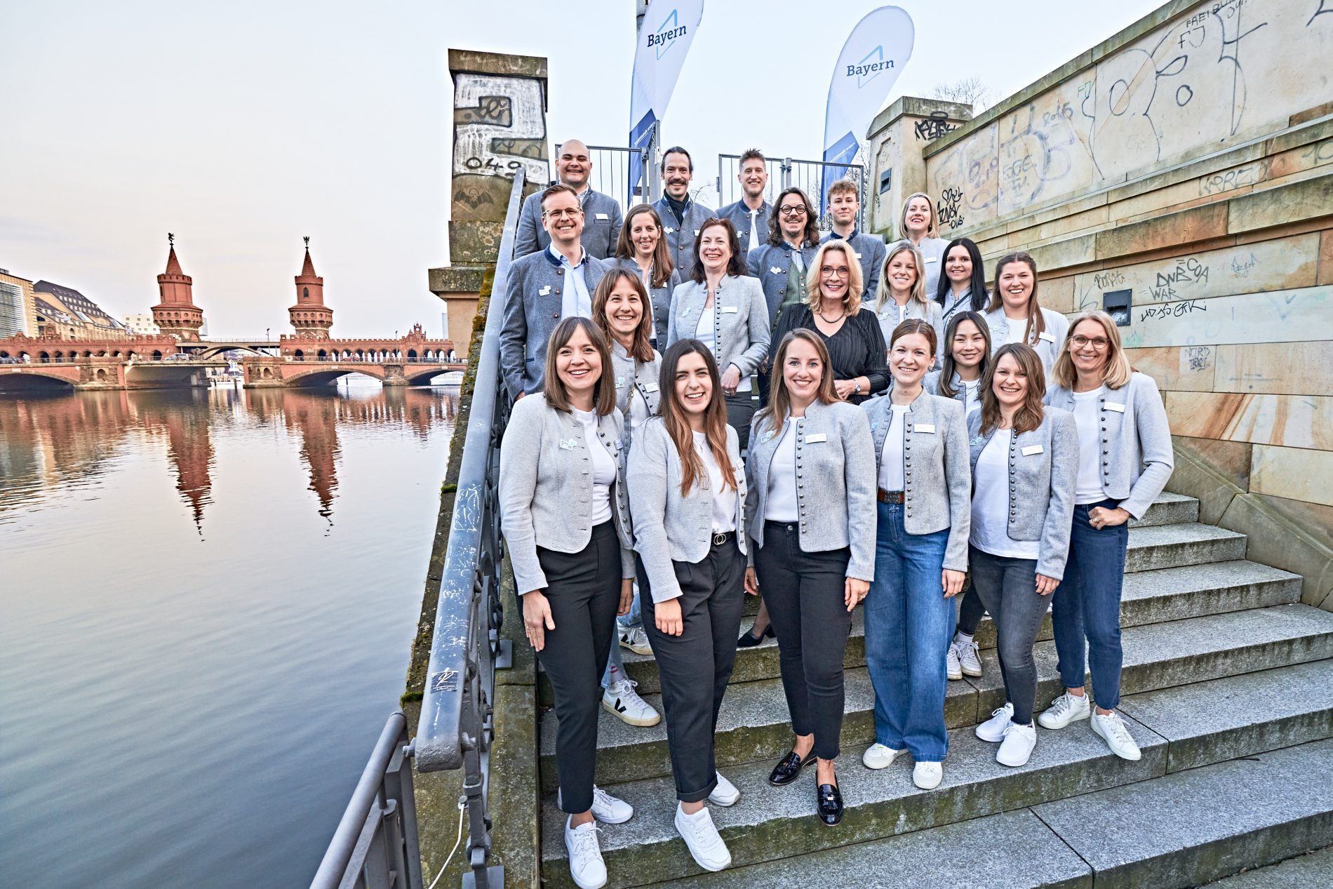 Bayern Connect meets Berlin Netzwerkevent Gruppe von ca. 20 Personen in grauen Blazern auf Treppen am Spreeufer; Bayern-Banner im Hintergrund (Event Bayern Connect meets Berlin 2024)