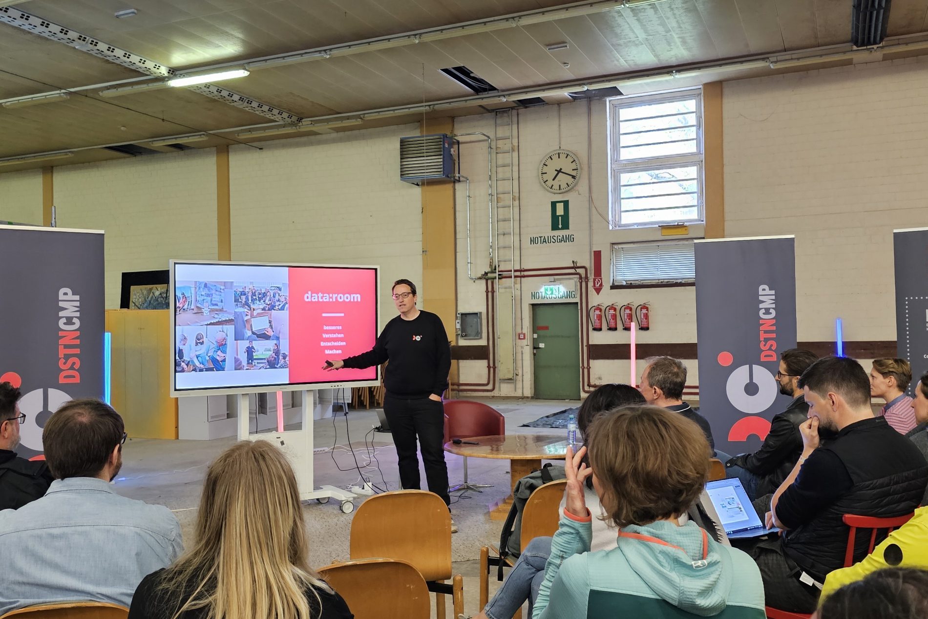 Referent präsentiert data:room-Folie vor Publikum in einer Industriehalle während DSTNCMP 2024.