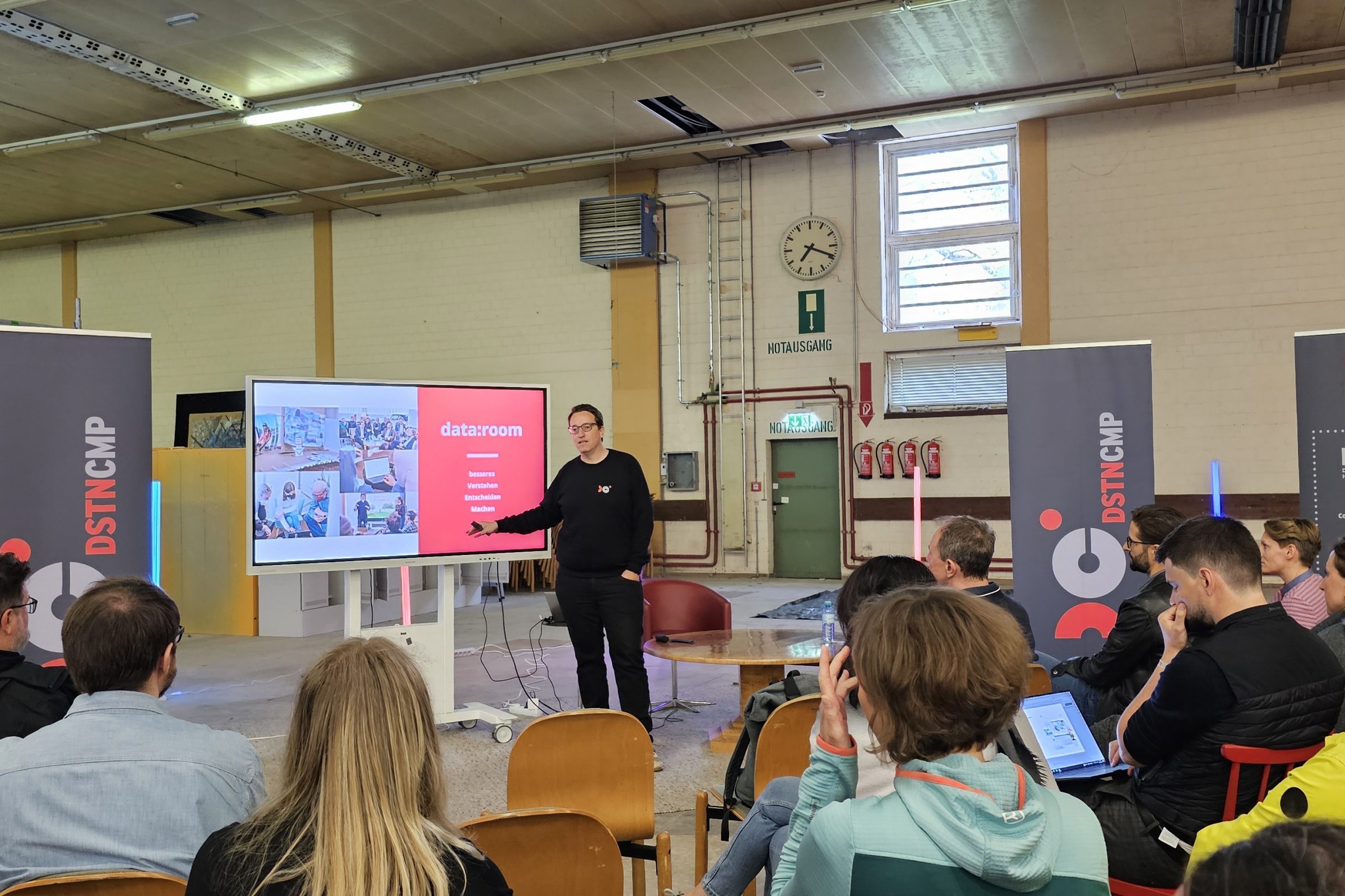 Referent pr&auml;sentiert data:room-Folie vor Publikum in einer Industriehalle w&auml;hrend DSTNCMP 2024.
