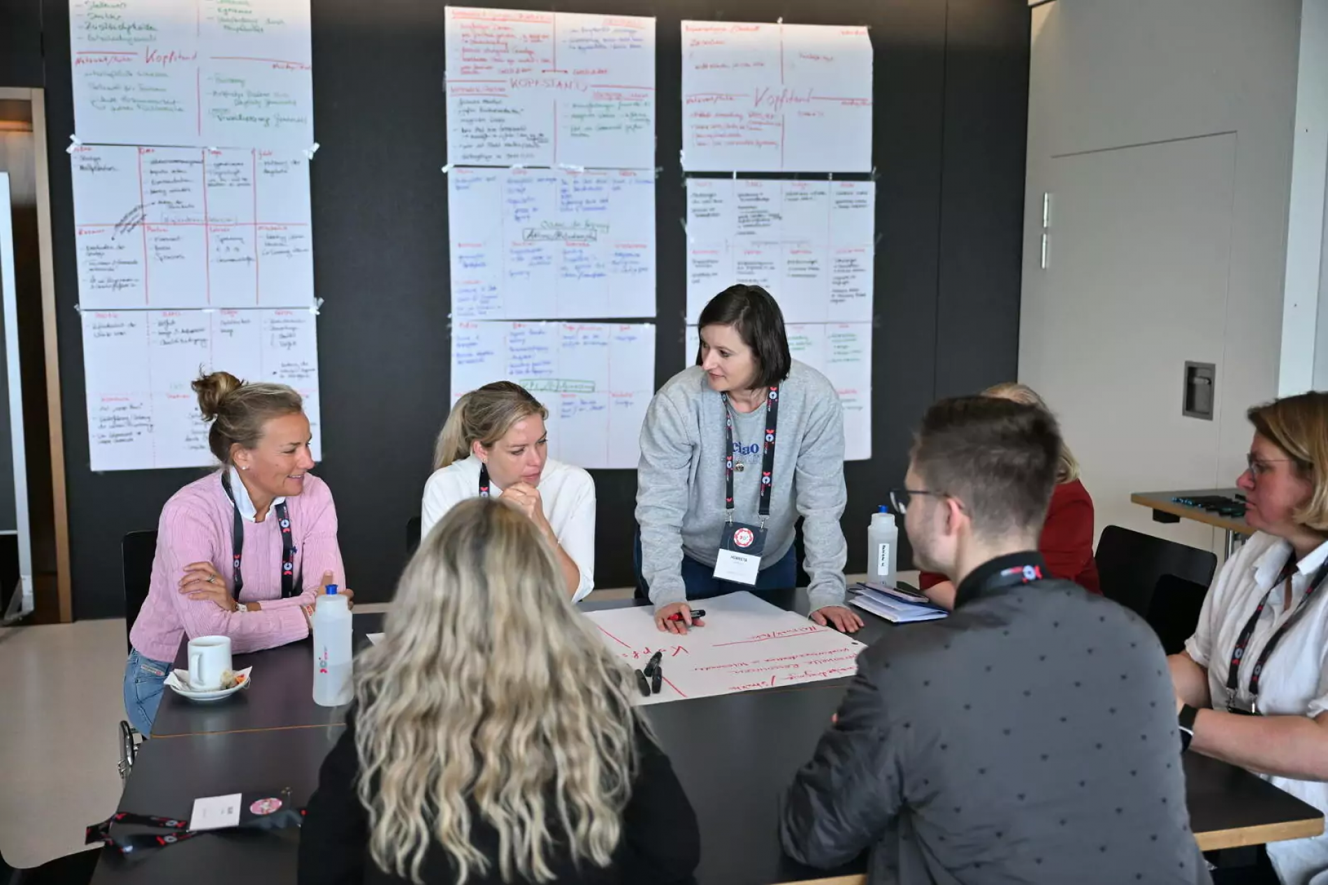 Teammitglieder diskutieren an einem rechteckigen Tisch; hinter ihnen große Pläne-Poster an der Wand.