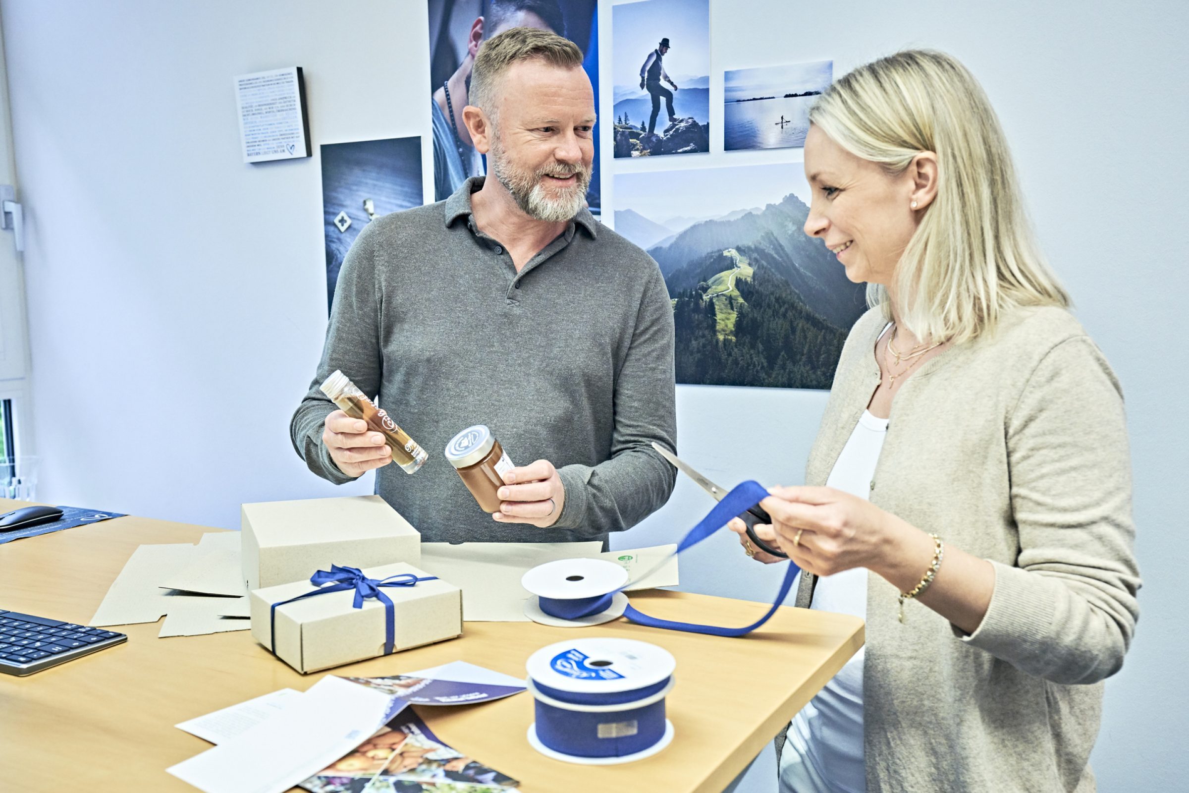 Zwei Mitarbeiter verpacken Geschenke am Schreibtisch; Geschenkboxen, Band, Schere, Rollband, Bergfotos im Hintergrund.