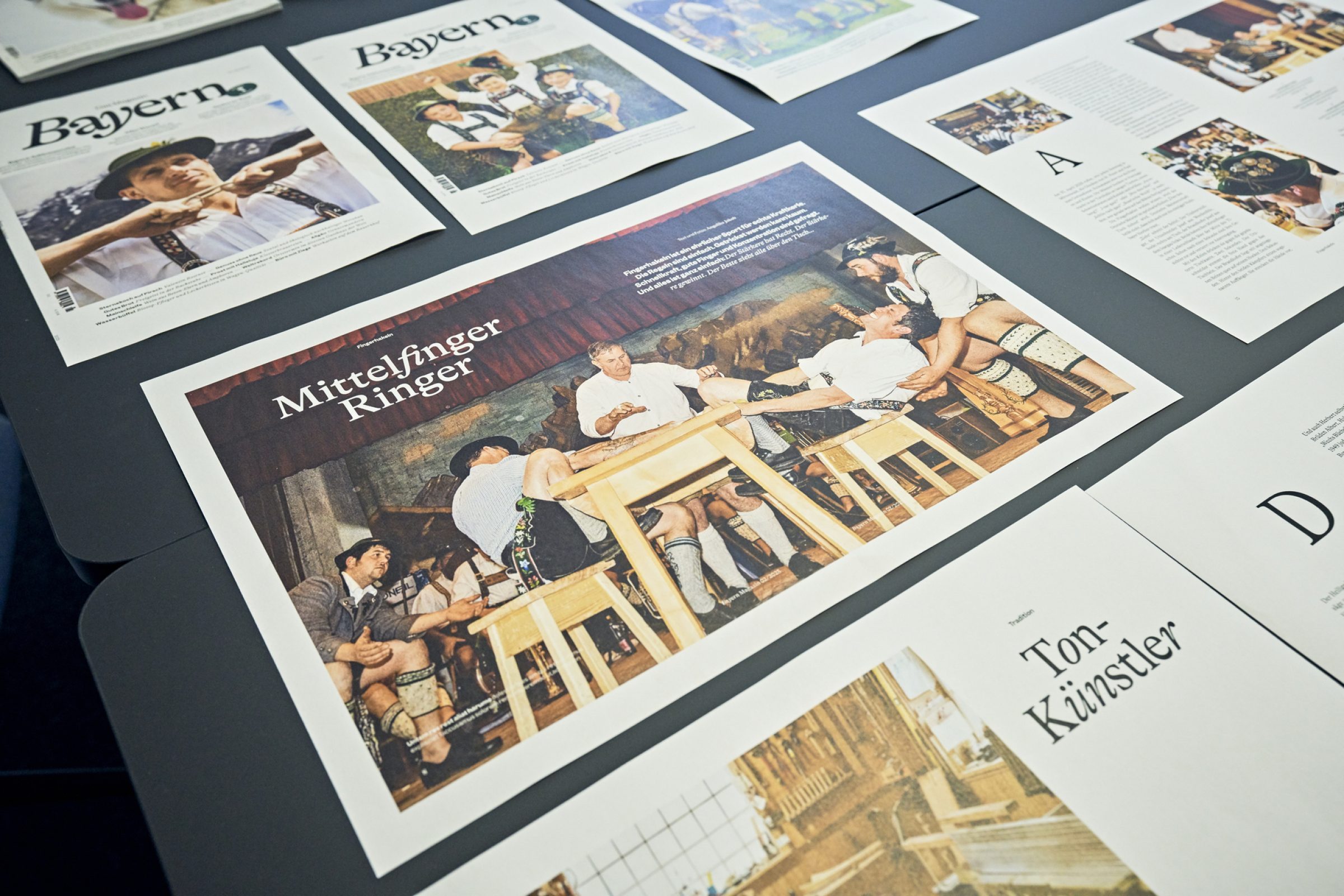 Mehrere BayTM Magazin-Cover liegen aus; im Vordergrund eine Wrestling-Szene mit Akteuren auf Holzb&auml;nken w&auml;hrend einer Show.