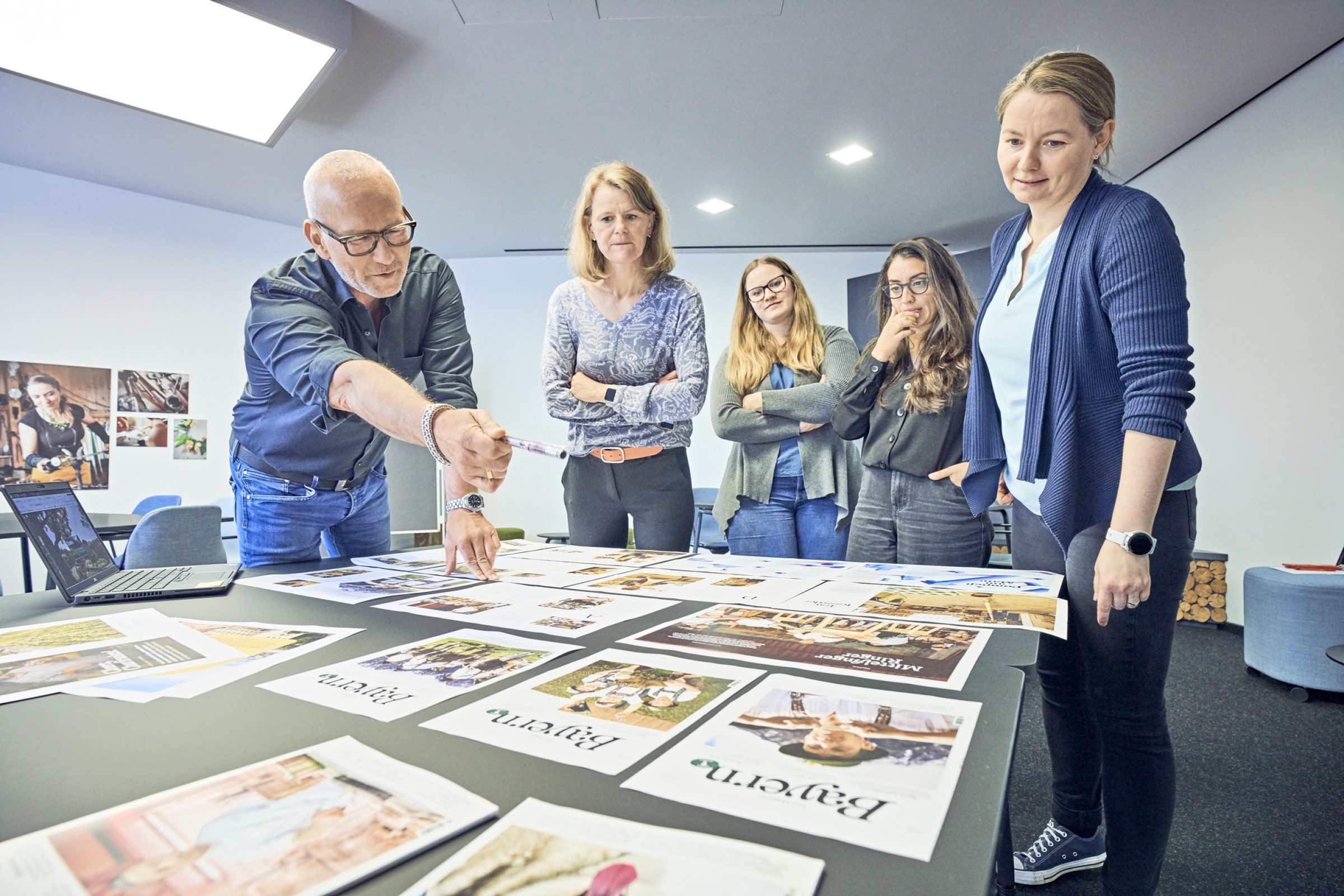F&uuml;nf Personen im Besprechungsraum pr&uuml;fen Marketingbrosch&uuml;ren und Layouts auf dem Tisch, Laptop offen.