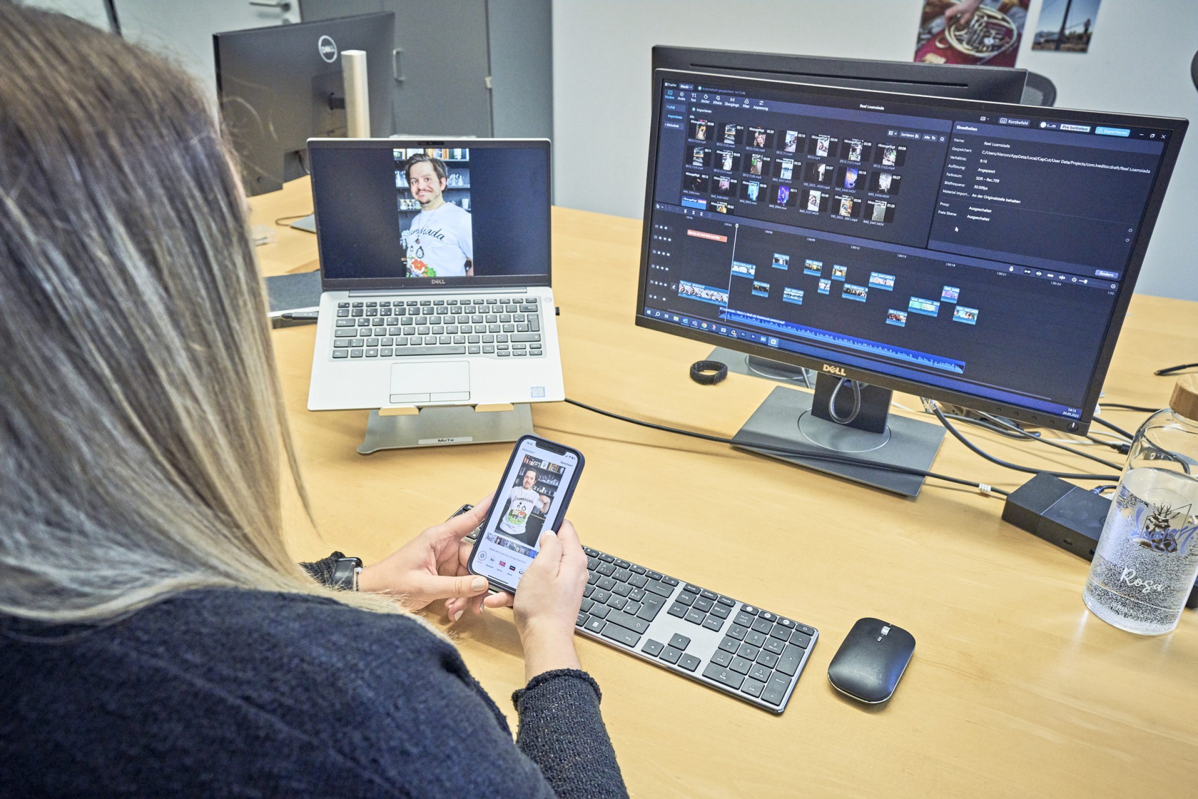 Person am Schreibtisch mit Laptop, externem Monitor und Smartphone; Laptop zeigt Foto, Monitor bearbeitet Videoprojekte.