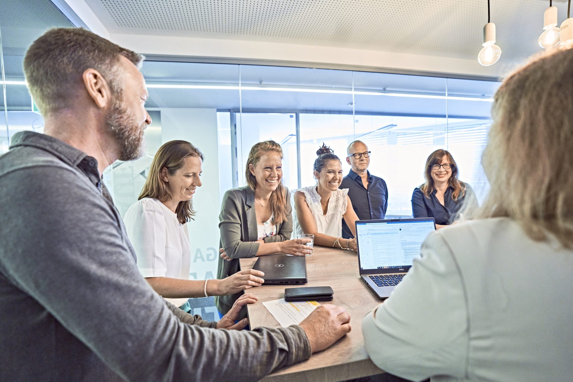 Ein Tag im Marketing der BayTM, Geschaeftsbericht 2023 BayTM Marketingtag: Team diskutiert um Konferenztisch, Laptop offen, Gläser Wasser, freundliche Gruppenarbeit.