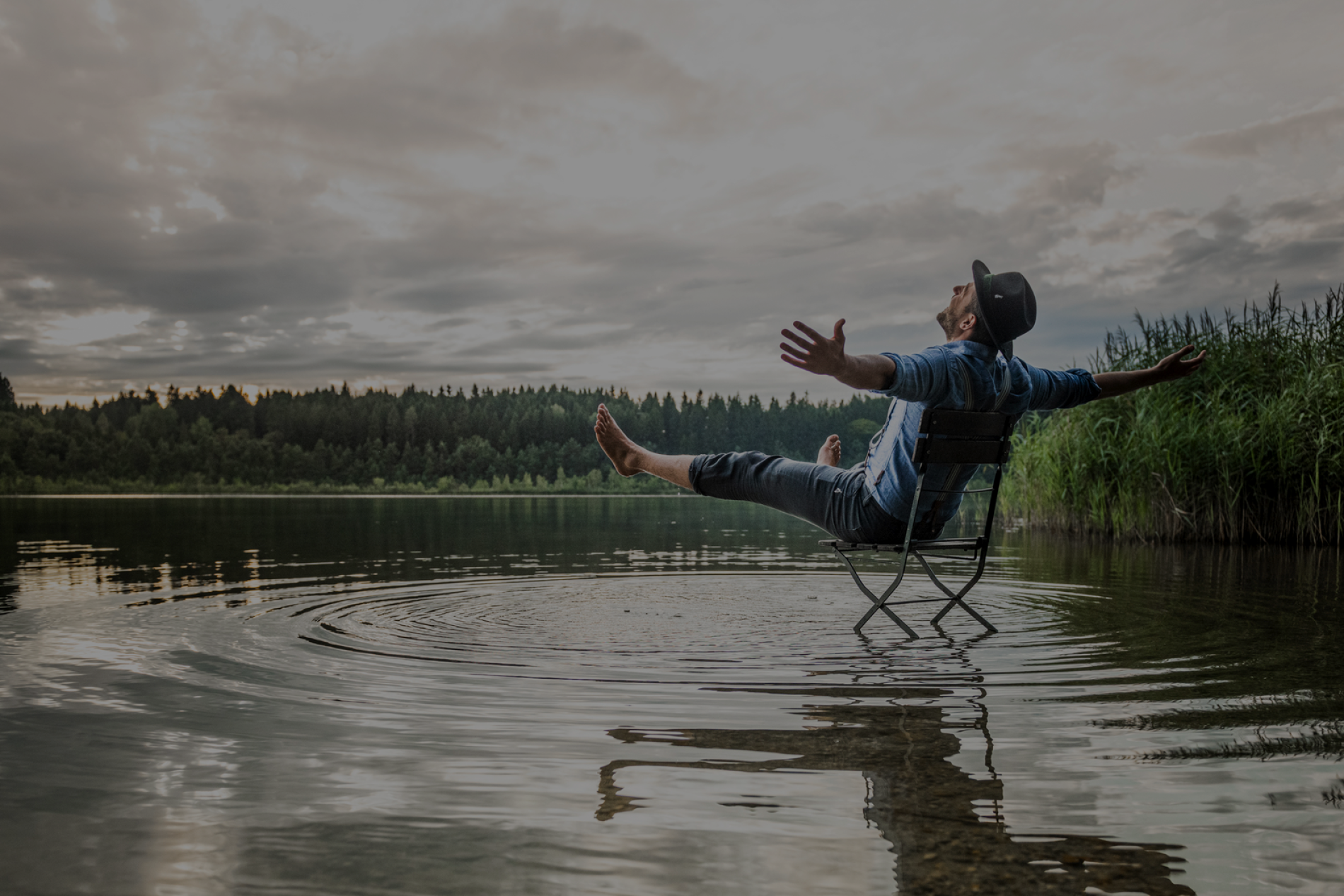077_8925263-JPG Website Breite_Width 1920px (300dpi)2 Mann sitzt in klappbarem Stuhl im Seewasser, Arme ausgebreitet, Hut, Wolkenhimmel, Ufergras am Rand.