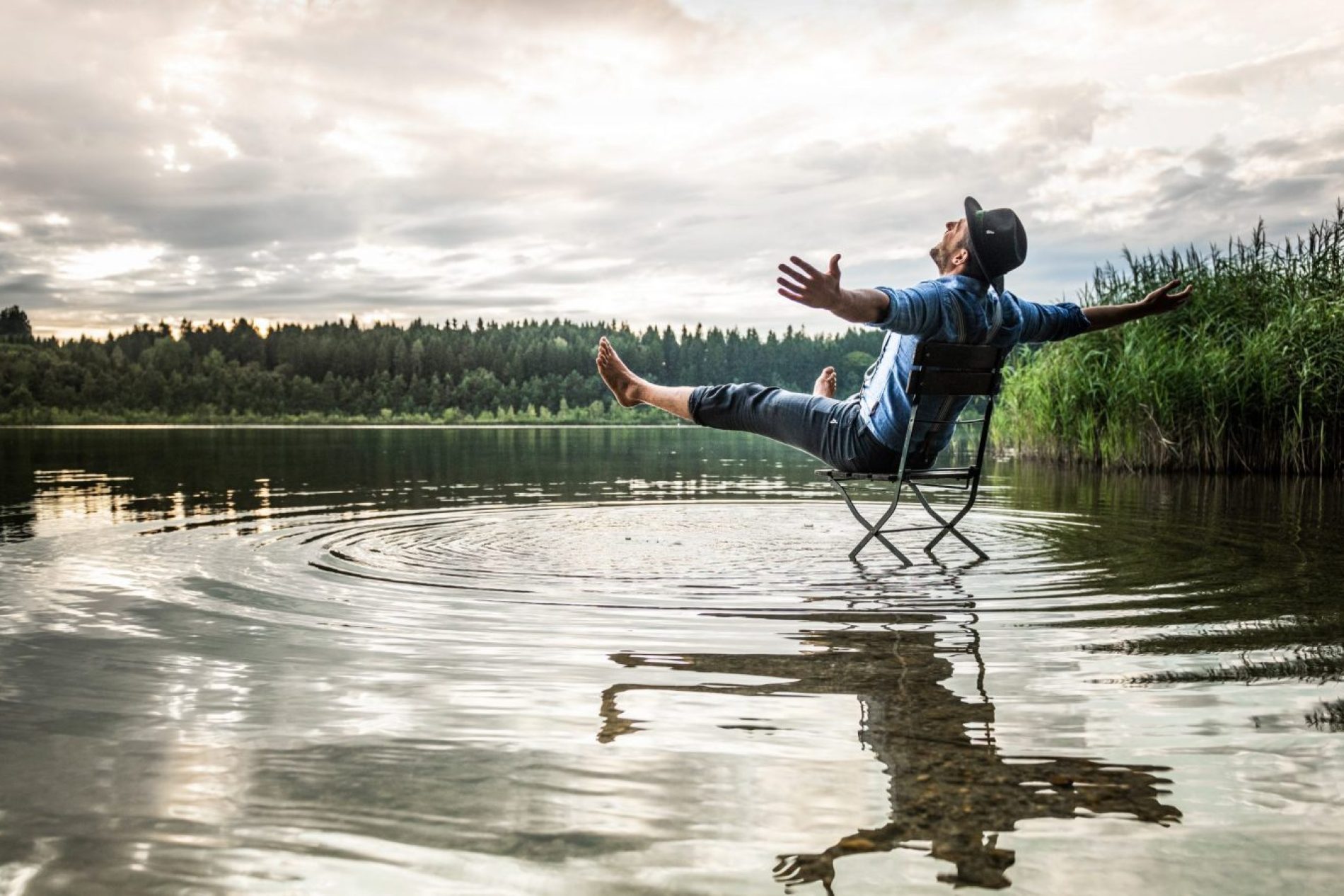 Mann sitzt in einem Klappstuhl im See, Arme ausgebreitet, Hut, Wolkenhimmel, Ufergras, Wasserreflexion.
