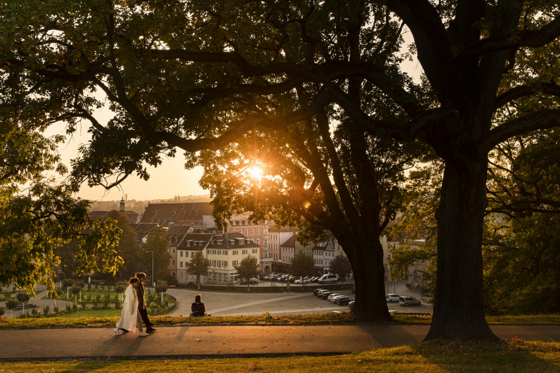 Paar geht bei Sonnenuntergang am Uferweg vorbei, im Vordergrund großer Baum; Stadt mit Mehrfamilienhäusern im Hintergrund.