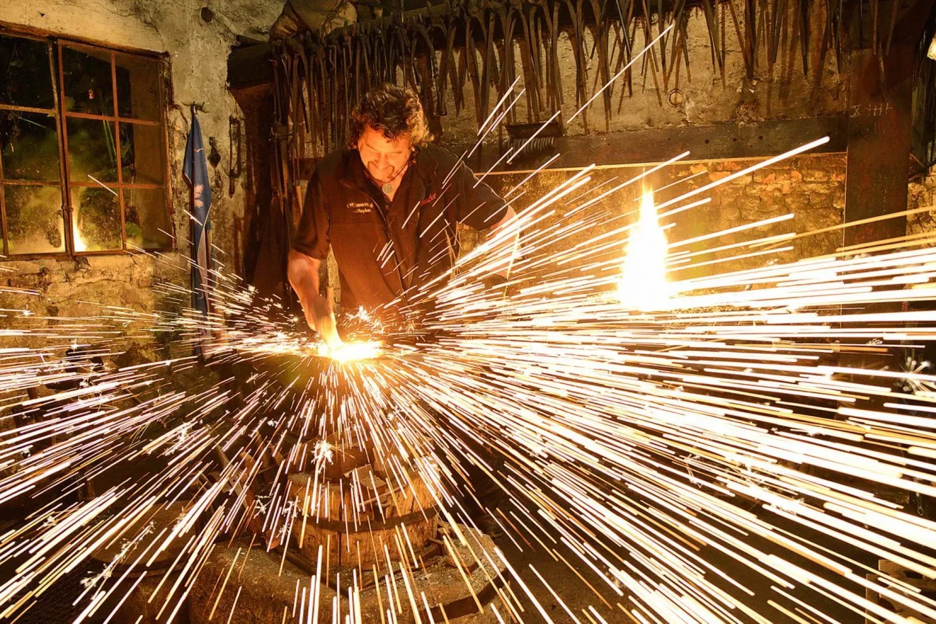 Schmied in dunkler Werkstatt bearbeitet Metall; Funken fliegen in alle Richtungen, Werkzeuge hängen an der Wand.