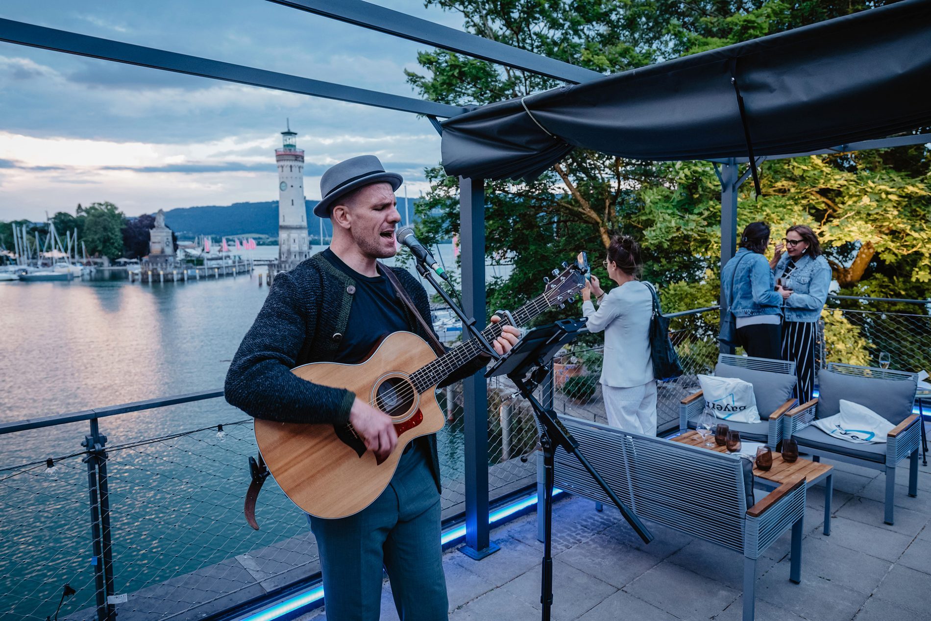 Deutschland, Bayern, Allgäu, Lindau, Bavaria Workshop 2024 Musiker singt mit Akustikgitarre am Bodensee während Bavaria Workshop; Blick auf Lindauer Leuchtturm, Gäste im Hintergrund.