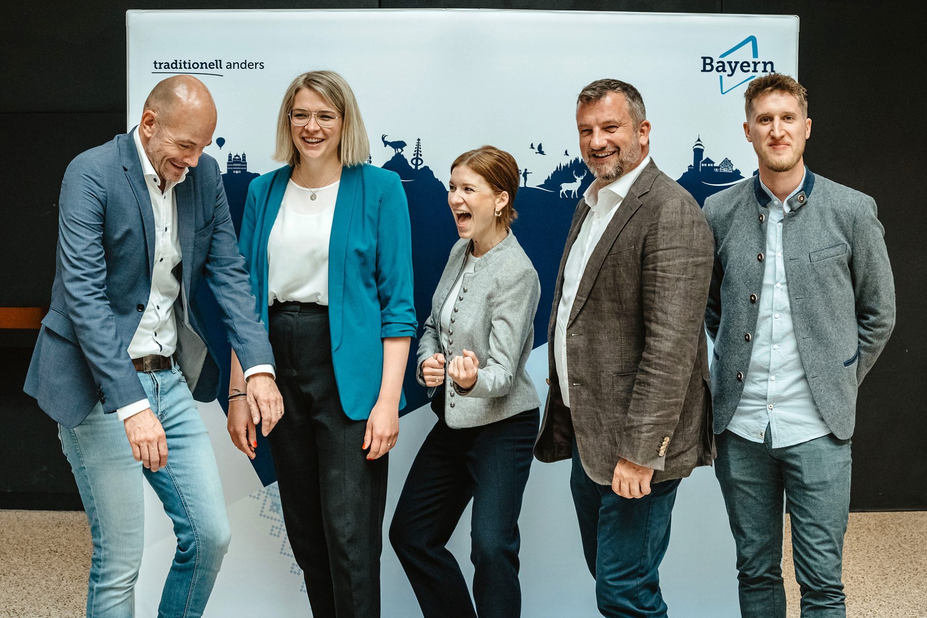 Deutschland, Bayern, Allgäu, Lindau, Bavaria Workshop 2024 Fünf Personen posieren vor Banner Bavaria Workshop; lächelnd, Business-Casual-Outfits, Allgäu-Silhouetten im Hintergrund.