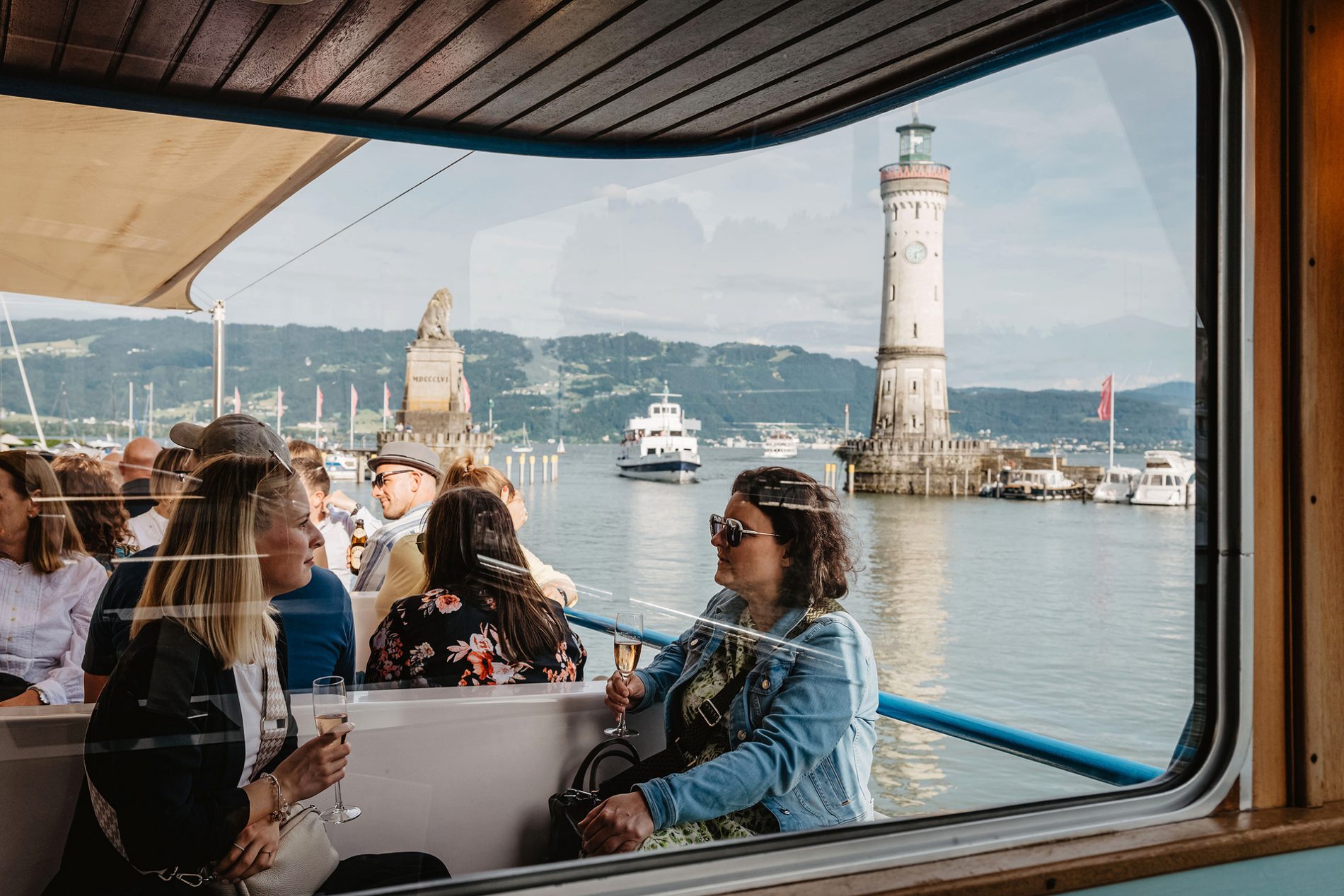 Deutschland, Bayern, Allgäu, Lindau, Bavaria Workshop 2024 Gruppe Passagiere auf einem Boot, Lindauer Leuchtturm und Hafen im Hintergrund, Gespräche mit Getränken.