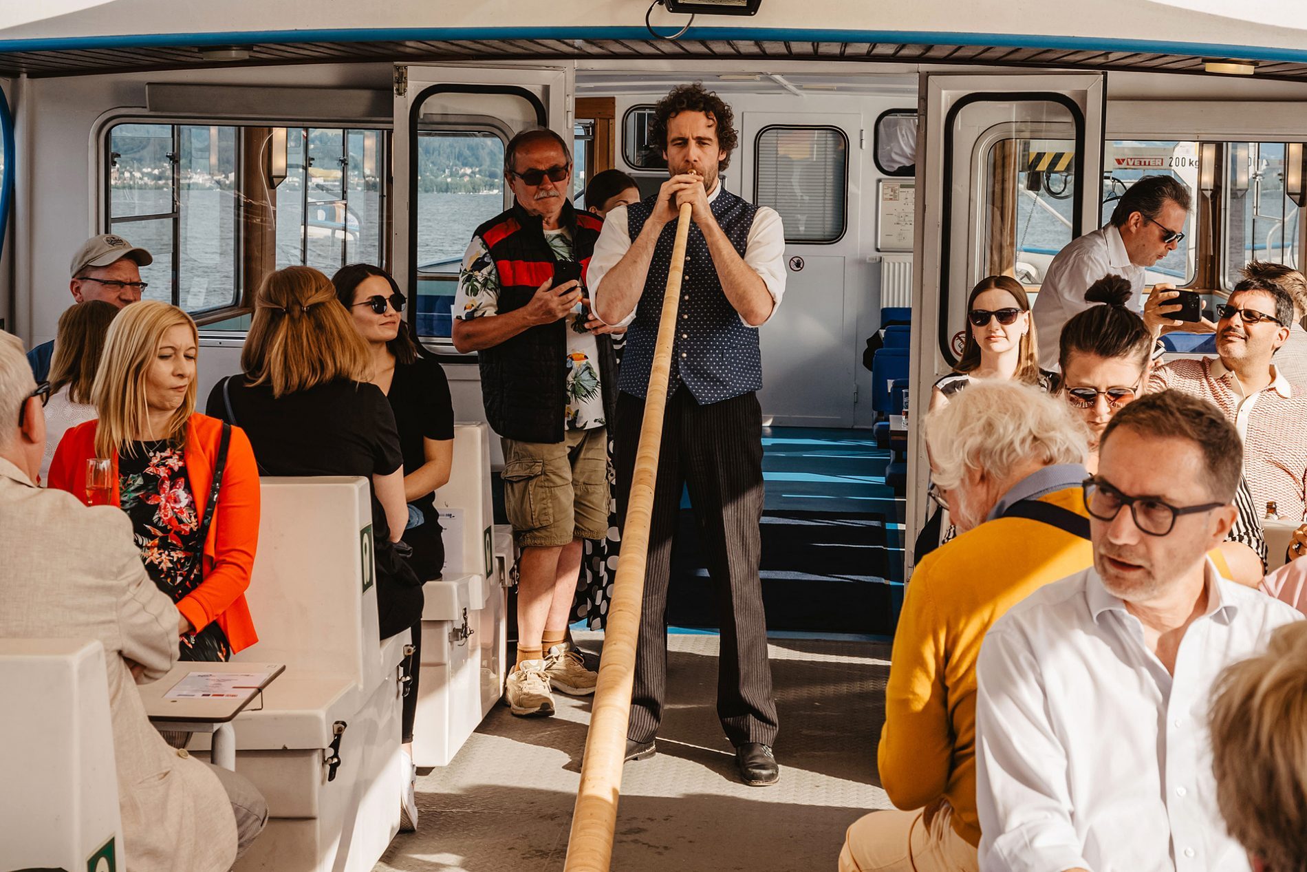 Deutschland, Bayern, Allgäu, Lindau, Bavaria Workshop 2024 Teilnehmer der Bavaria Workshop sitzen in einem Boot; ein Mann spielt ein Alphorn, Publikum hört zu.