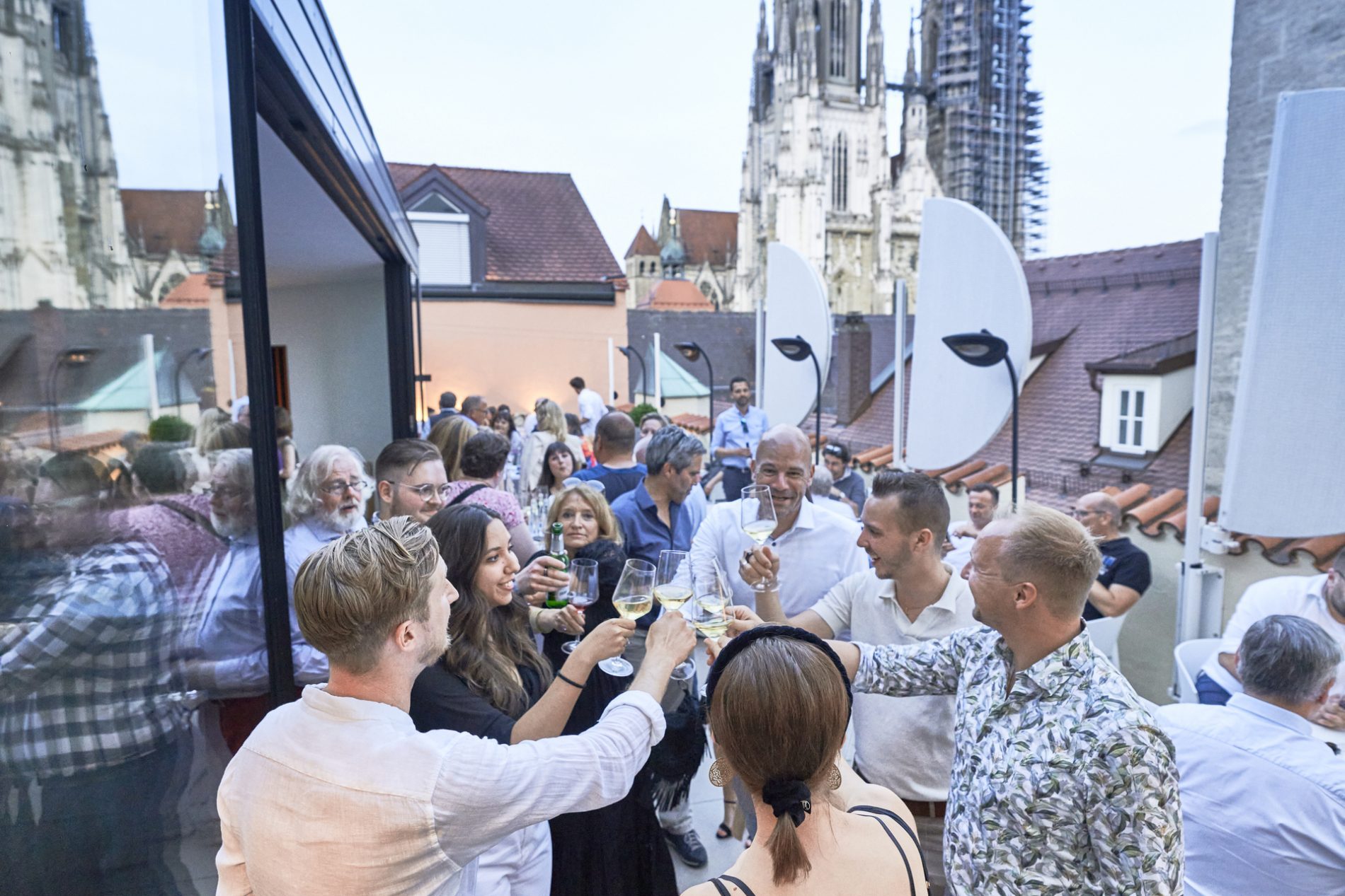 Bavaria Workshop Regensburg, Abendevent im Restaurant „Storstad“ Bavaria Workshop Abend in Regensburg auf Dachterrasse Storstad; Gäste stoßen mit Wein an, Kathedrale im Hintergrund.