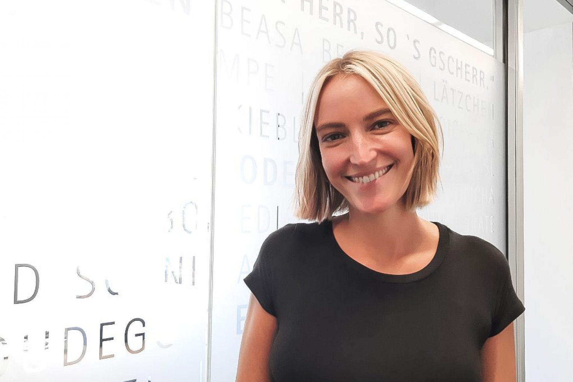 Luisa von Podewils Porträt von Luisa von Podewils, lächelnde Frau mit kurzen blonden Haaren in schwarzem Oberteil vor Glaswand mit Schriftzug.