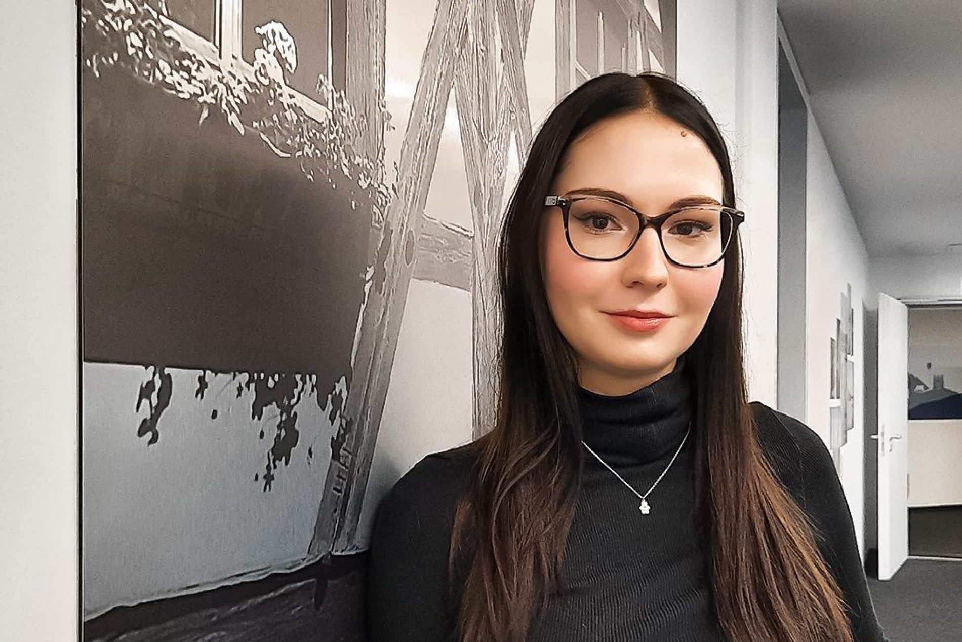 Marisa_1020x765 Porträt einer jungen Frau mit Brille, schwarzem Rollkragenpullover und Halskette vor einem Schwarz-Weiß-Wandbild im Büroflur.