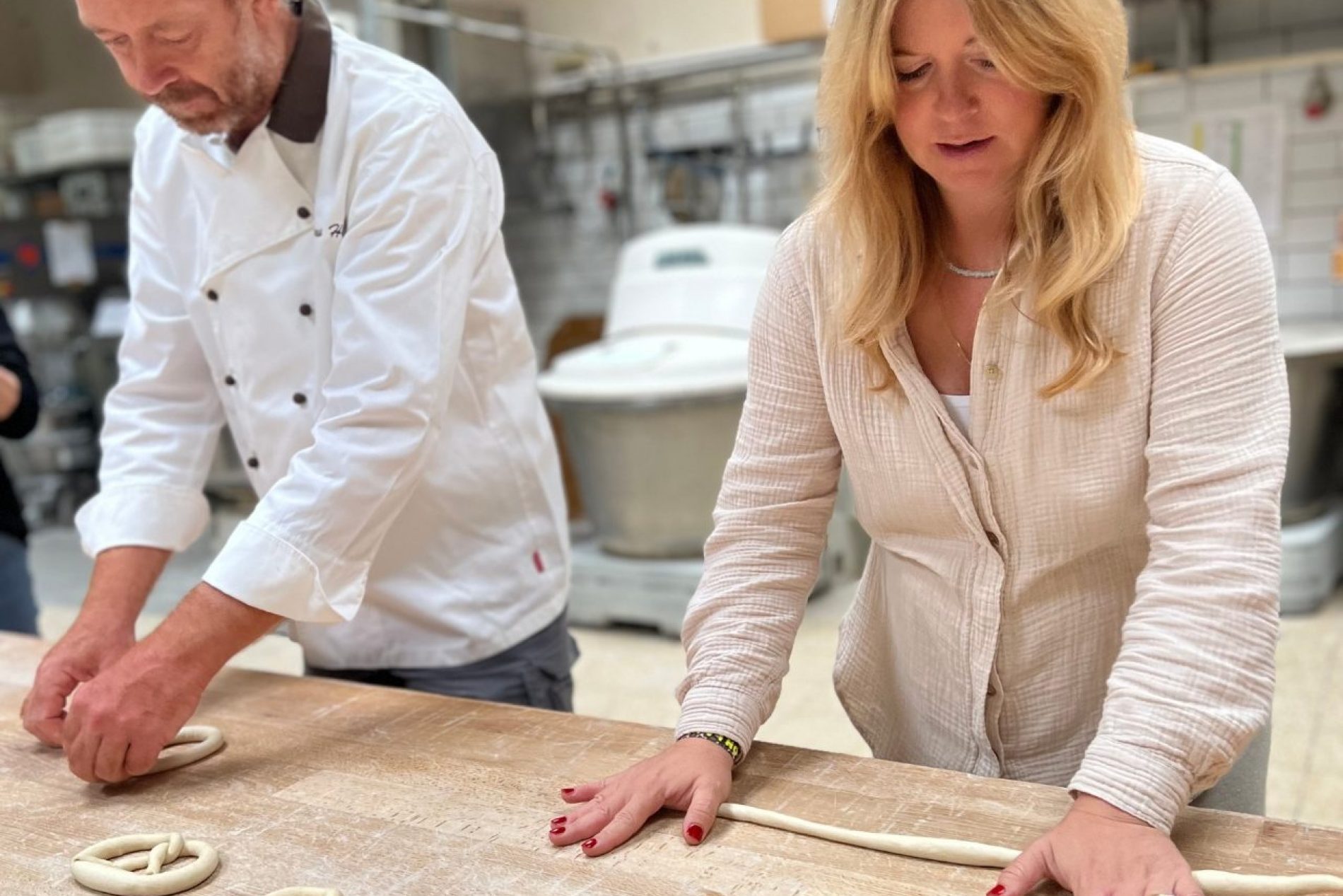 Eine Mann und ein Frau stehen an einem Holztisch und formen Teig zu Brezeln.