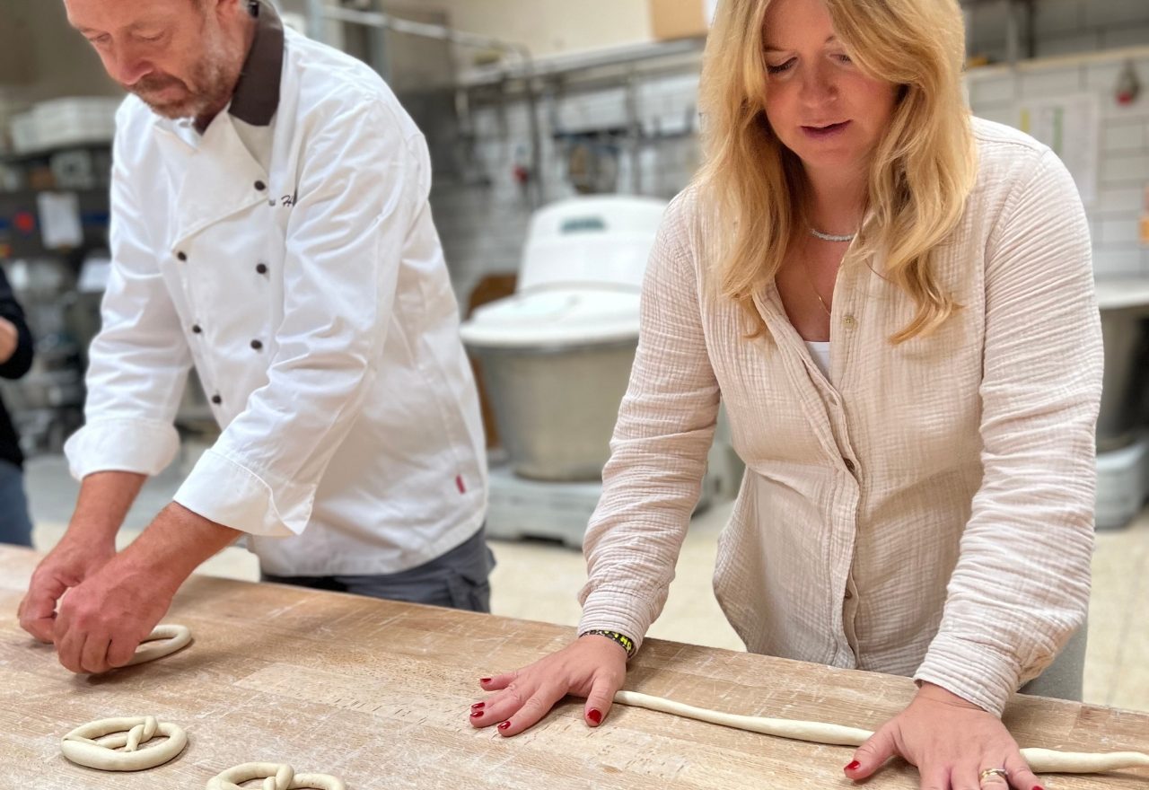 Eine Mann und ein Frau stehen an einem Holztisch und formen Teig zu Brezeln.